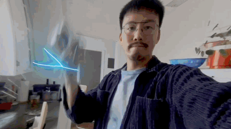 A young man with glasses taking a selfie in a kitchen, holding a glowing electronic device that creates visual light streaks.