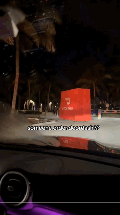 Nighttime scene viewed from inside a car showing a red DoorDash sign and a few people near a sidewalk, with trees and a building in the background.