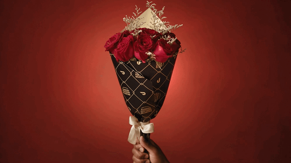 Hand holding a bouquet of red roses wrapped in black paper with gold geometric patterns and a white ribbon, against a red background.