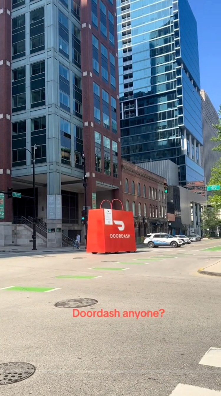 A large red DoorDash delivery bag placed on the street in front of tall modern office buildings, with cars parked nearby and a person riding a bicycle, and a green traffic light at the intersection.