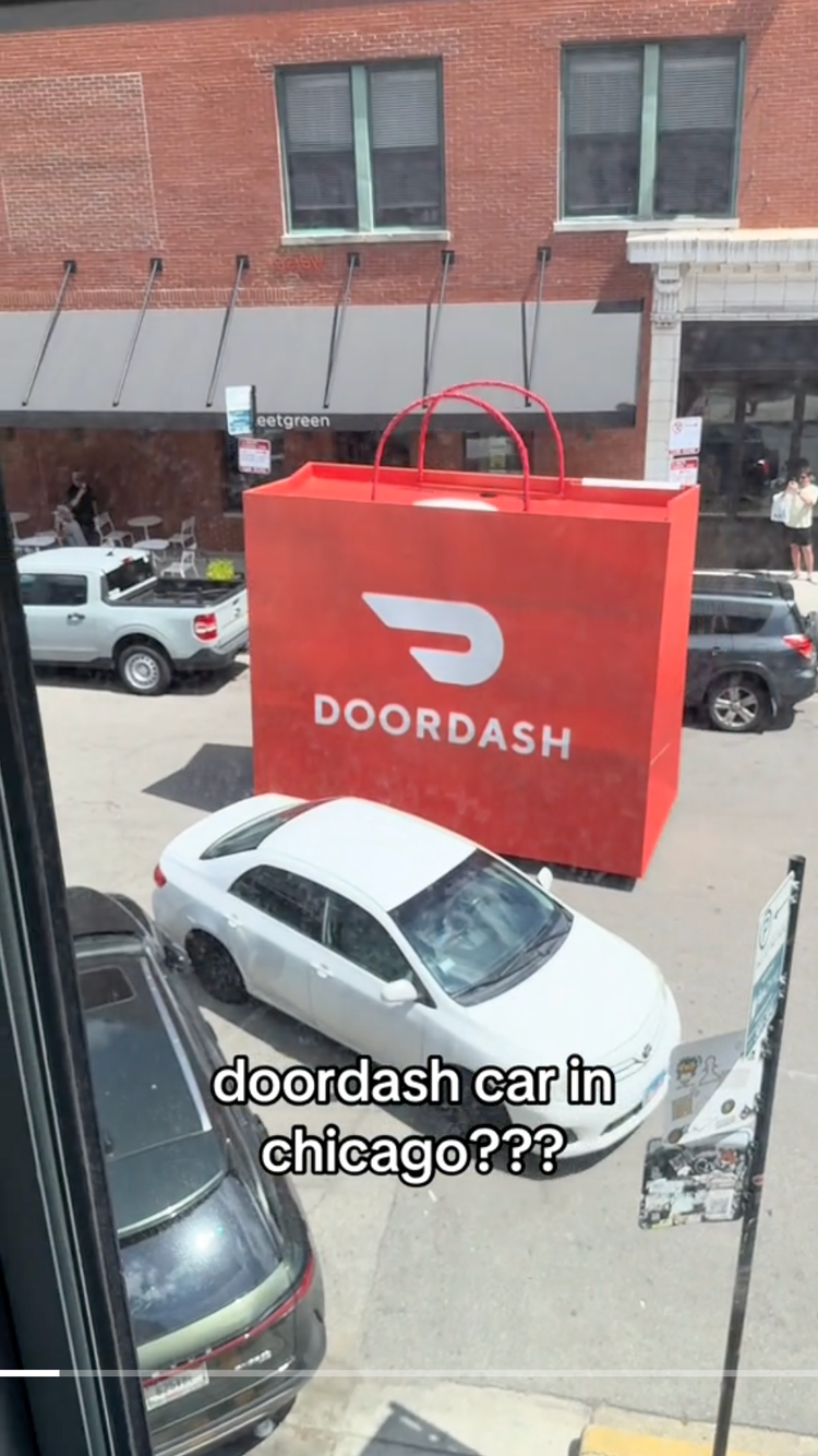 Large red shopping bag with the DoorDash logo hanging outside a store in Chicago, with parked cars and a brick building in the background.