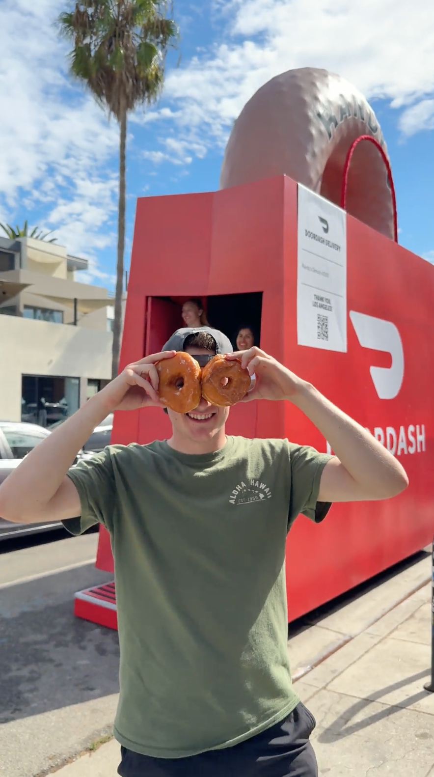Person holding two glazed donuts in front of their eyes, smiling, standing outdoors near a large red and pink Dunkin' Donuts sign and building, with a partly cloudy sky and palm trees in the background.