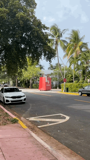 Street scene with cars, palm trees, and a large red mailbox or structure in the background.