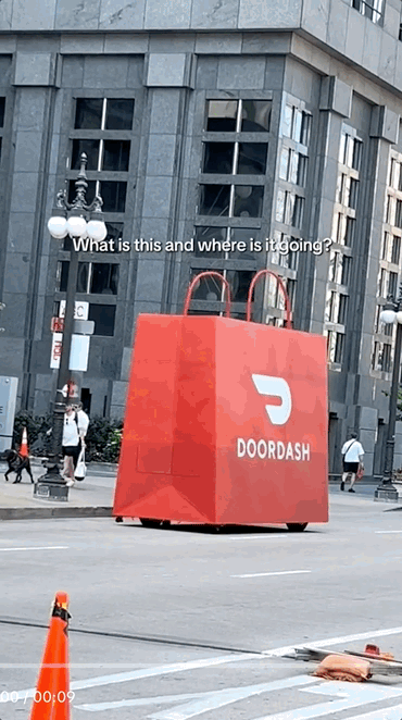 A large red DoorDash delivery bag shaped like a giant shopping bag on a city street, with a building and people walking in the background.