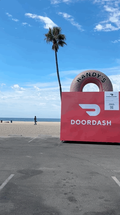 A large red DOORDASH sign with a donut-shaped RANDY'S sign behind it on a beach parking lot, with a palm tree, blue sky, ocean, and a person walking in the background.