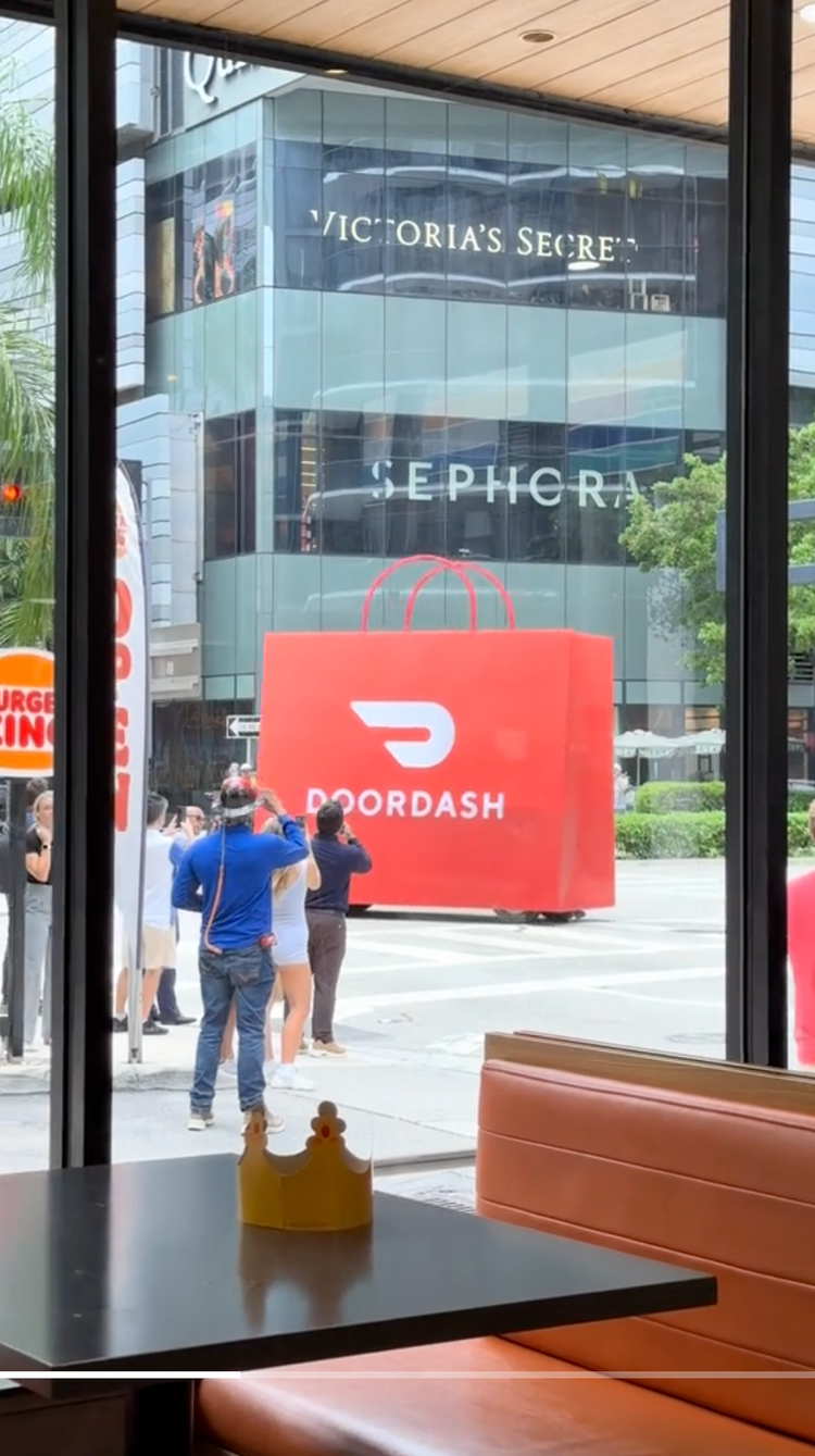 Photo taken from inside a restaurant showing a large red DoorDash advertisement on the window outside. Through the window, people are waiting in line outside, and the reflection of retail stores like Victoria's Secret and Sephora can be seen on the b