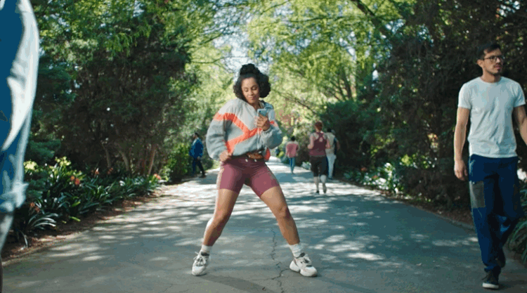 A woman dancing on a paved park pathway surrounded by trees, with other people walking in the background.