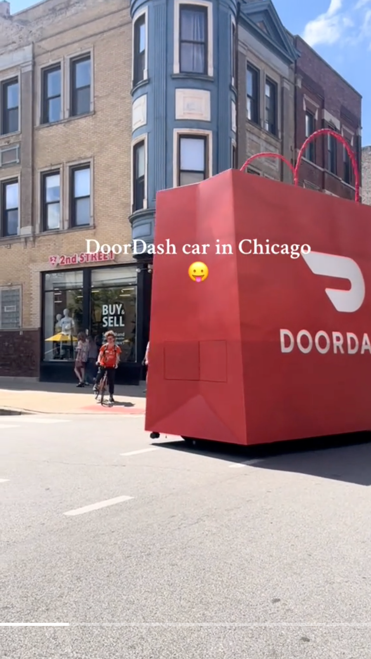A red DoorDash delivery car parked on a street in Chicago with historic brick buildings and people walking nearby.