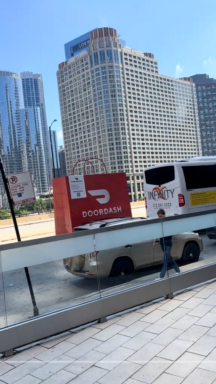 Urban scene with tall office buildings, a DOORDASH sign, a bus with 'INEQUALITY' branding, a tan car, and pedestrians walking on the sidewalk. Clear blue sky.