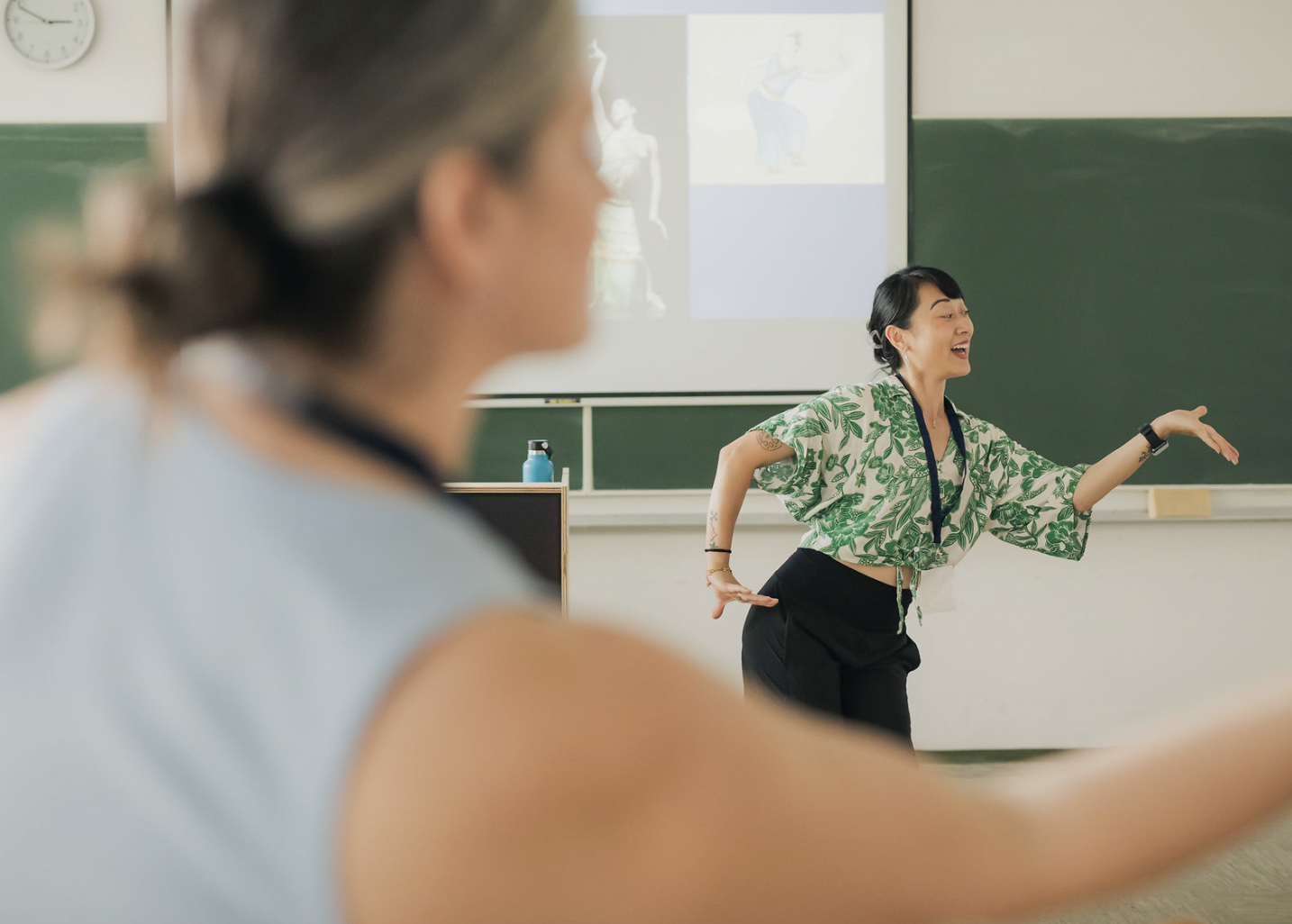 A woman in a green patterned blouse is standing in front of a classroom, gesturing with her right hand and smiling as she teaches students. A student with blurred features is in the foreground, and a presentation with images of dancers is projected on a screen behind her.