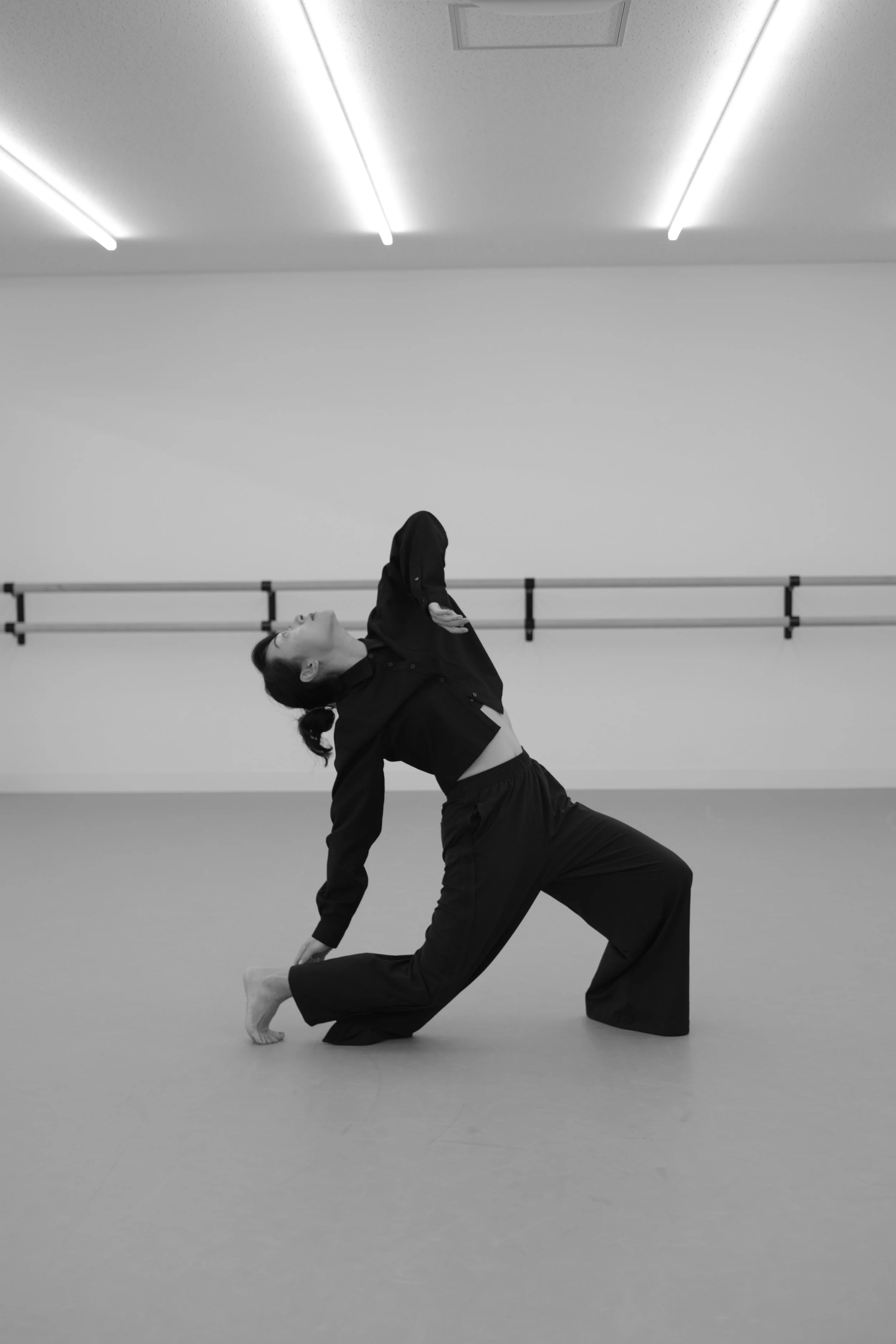 A woman in black clothing performs a dance or yoga pose in a minimalistic, brightly lit dance studio with a barre on the wall.