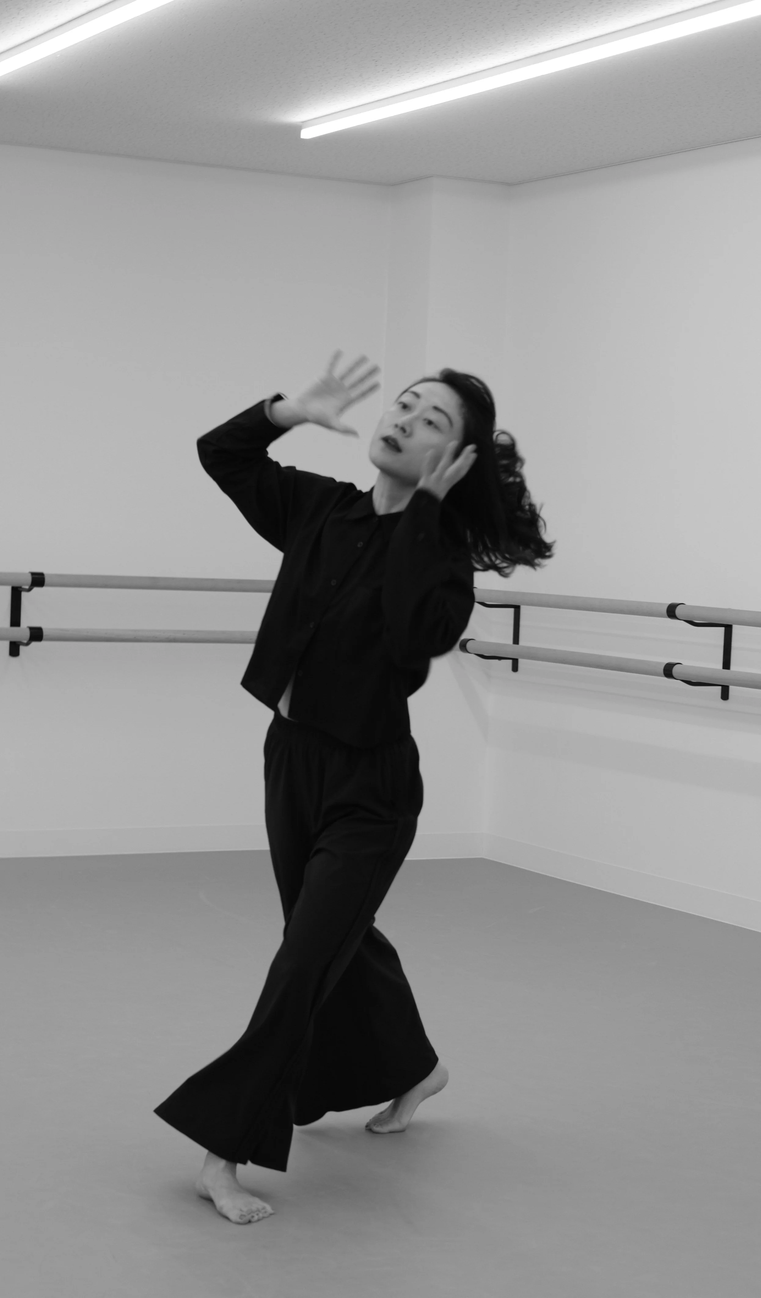 A person with dark hair dancing in a dance studio, wearing a black shirt and loose black pants, with a ballet barre in the background.