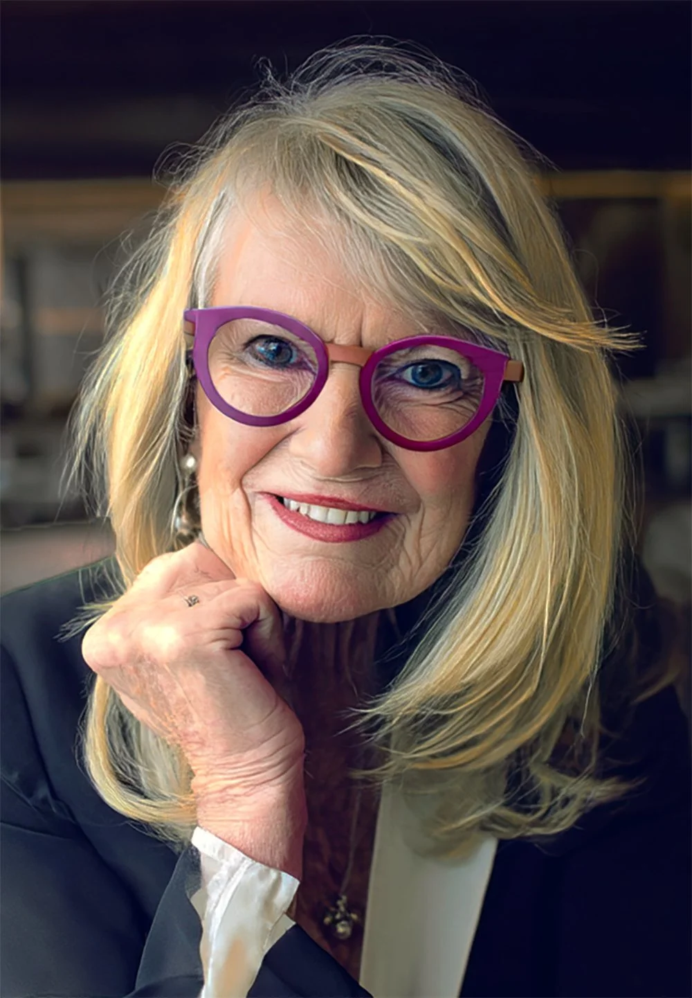 Close-up of an elderly woman with long blonde hair, wearing purple glasses, smiling, and resting her chin on her hand.