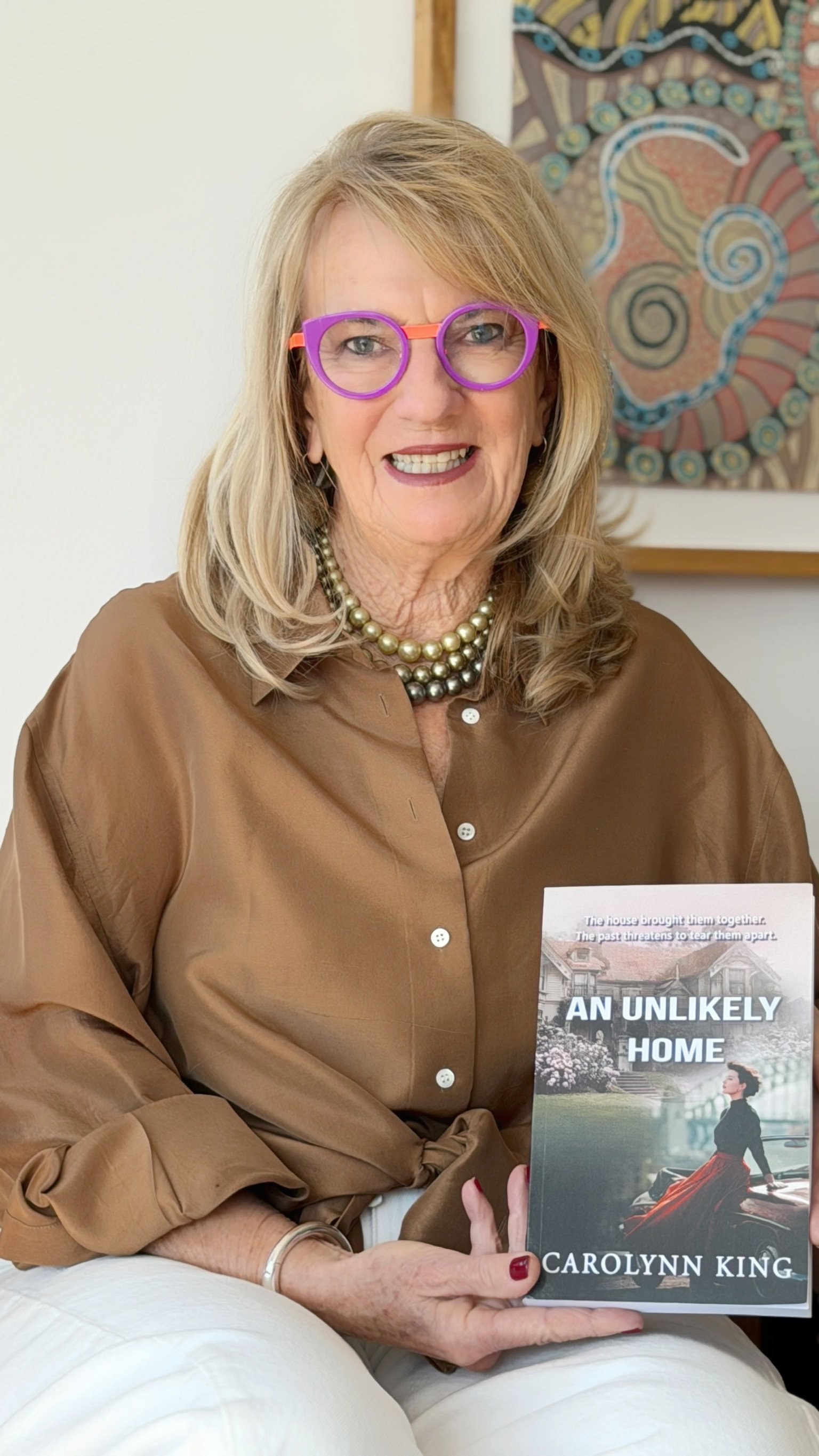 A woman sitting on a white chair holding a book titled "An Unlikely Home" by Carolynn King. She is wearing a brown satin blouse, a pearl necklace, and purple glasses with orange accents. She is smiling and has shoulder-length blonde hair. A colorful abstract painting is visible in the background.