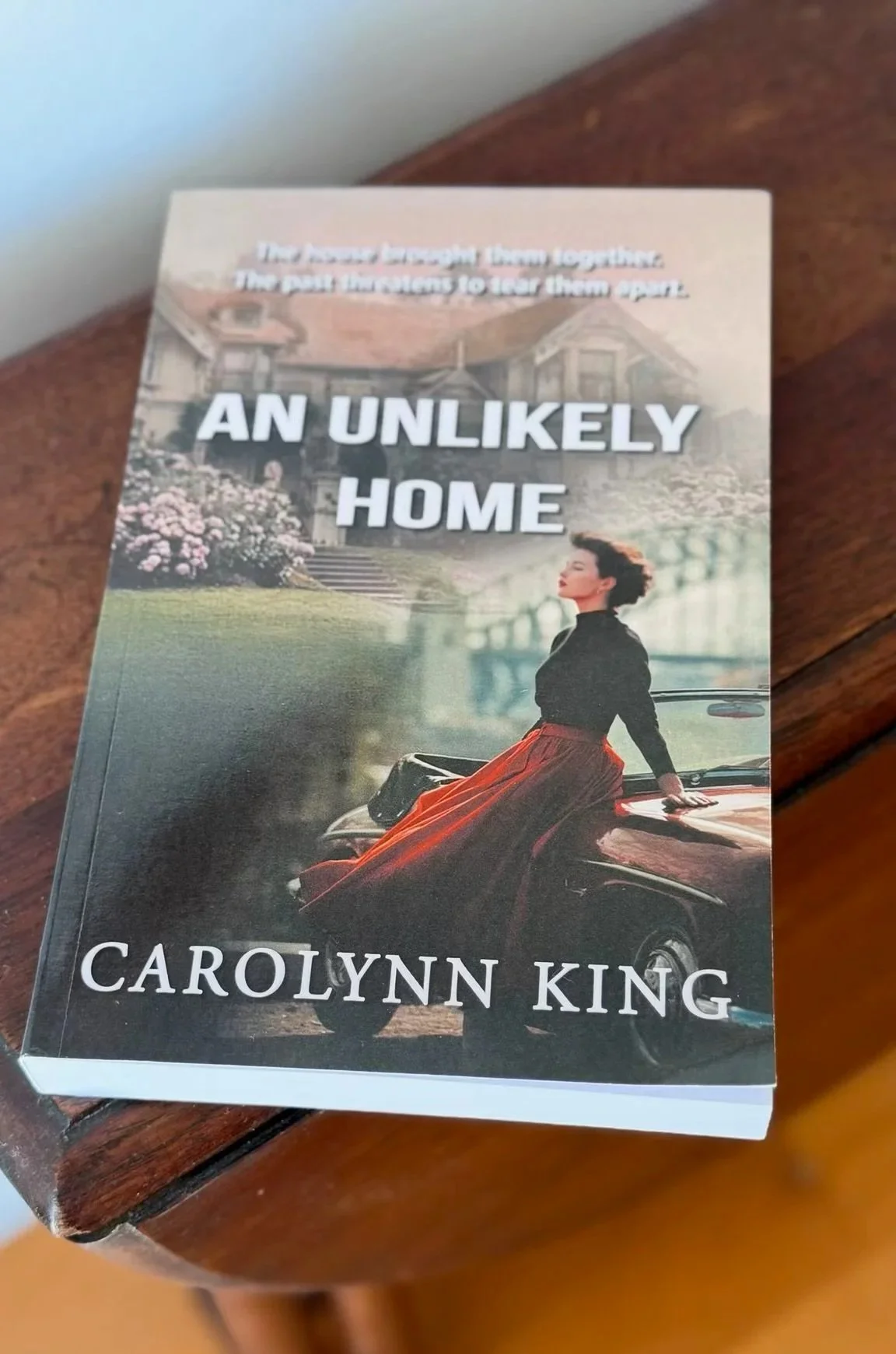 Book titled 'An Unlikely Home' by Carolynn King, featuring a woman in a black top and red skirt sitting on the hood of a vintage car in front of a house and garden.