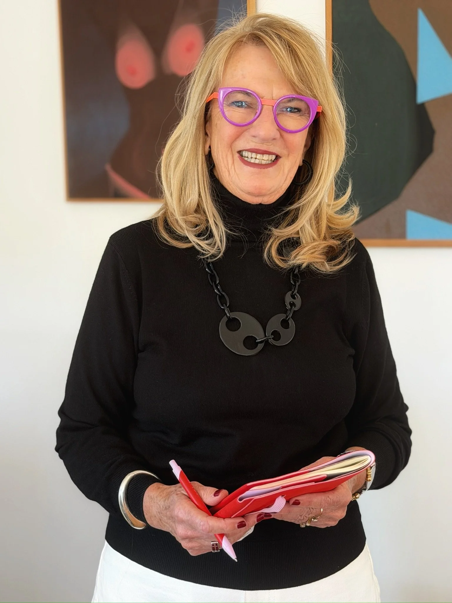 A woman with shoulder-length blonde hair wearing purple glasses and a black turtleneck sweater, holding a pink notebook and a red pen, standing in front of abstract artwork.