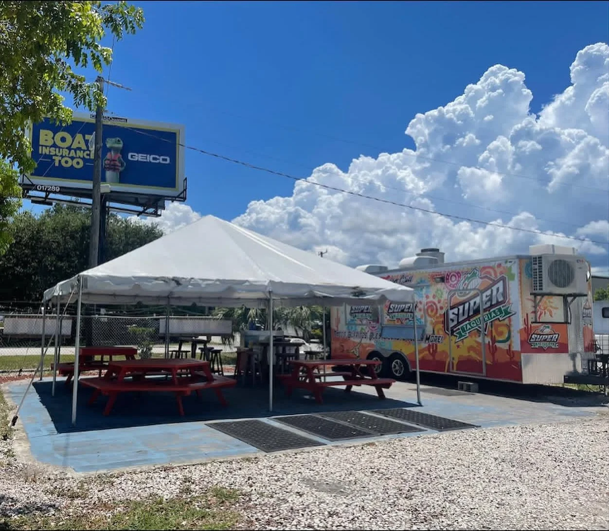 A food truck with colorful artwork and branding parked next to a white tent with picnic tables underneath, set on a concrete pad outside on a sunny day with blue sky and white clouds. A billboard advertising boat insurance is visible in the background.