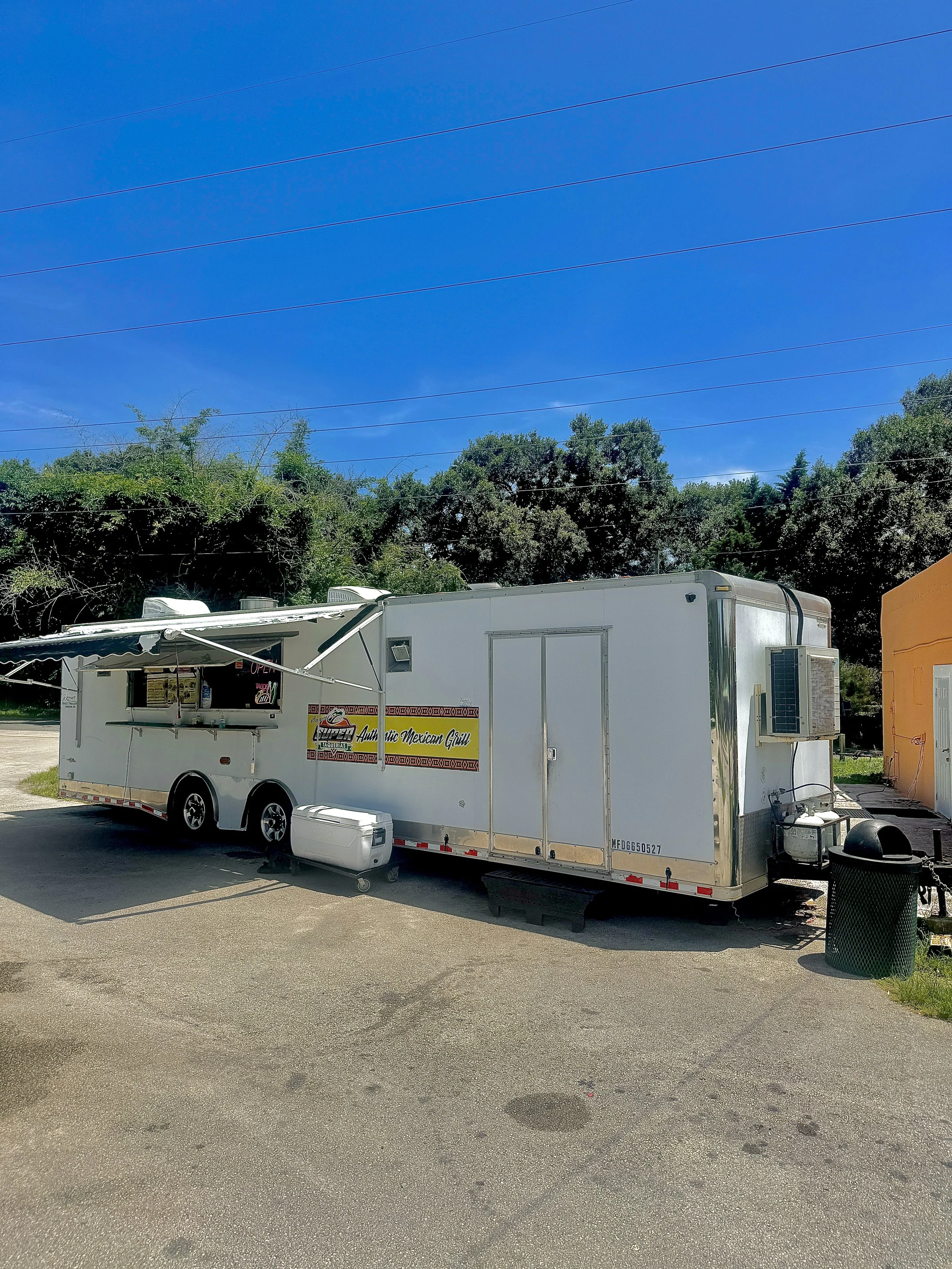 A white food truck offering authentic Mexican grilled food, parked on a paved lot with trees and a clear blue sky in the background. There is a black trash can and some outdoor equipment near the truck.