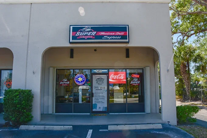 Front of a restaurant with a sign that reads 'Super Taqueria Mexican Grill Express'. The glass door has a menu and signs, including a Coca-Cola sign and an 'Open' sign. Parking spaces are visible in front.