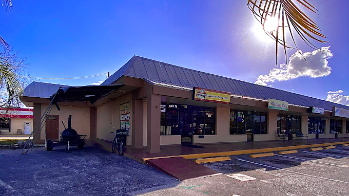 Single-story shopping plaza with several storefronts and large windows, a parking lot with yellow-striped parking space, under a bright blue sky with a cloud and palm leaves overhead.