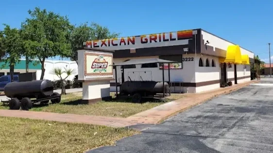 Exterior view of a Mexican grill restaurant with yellow awnings, a sign, and a small outdoor seating area, in a parking lot with trees and clear sky.