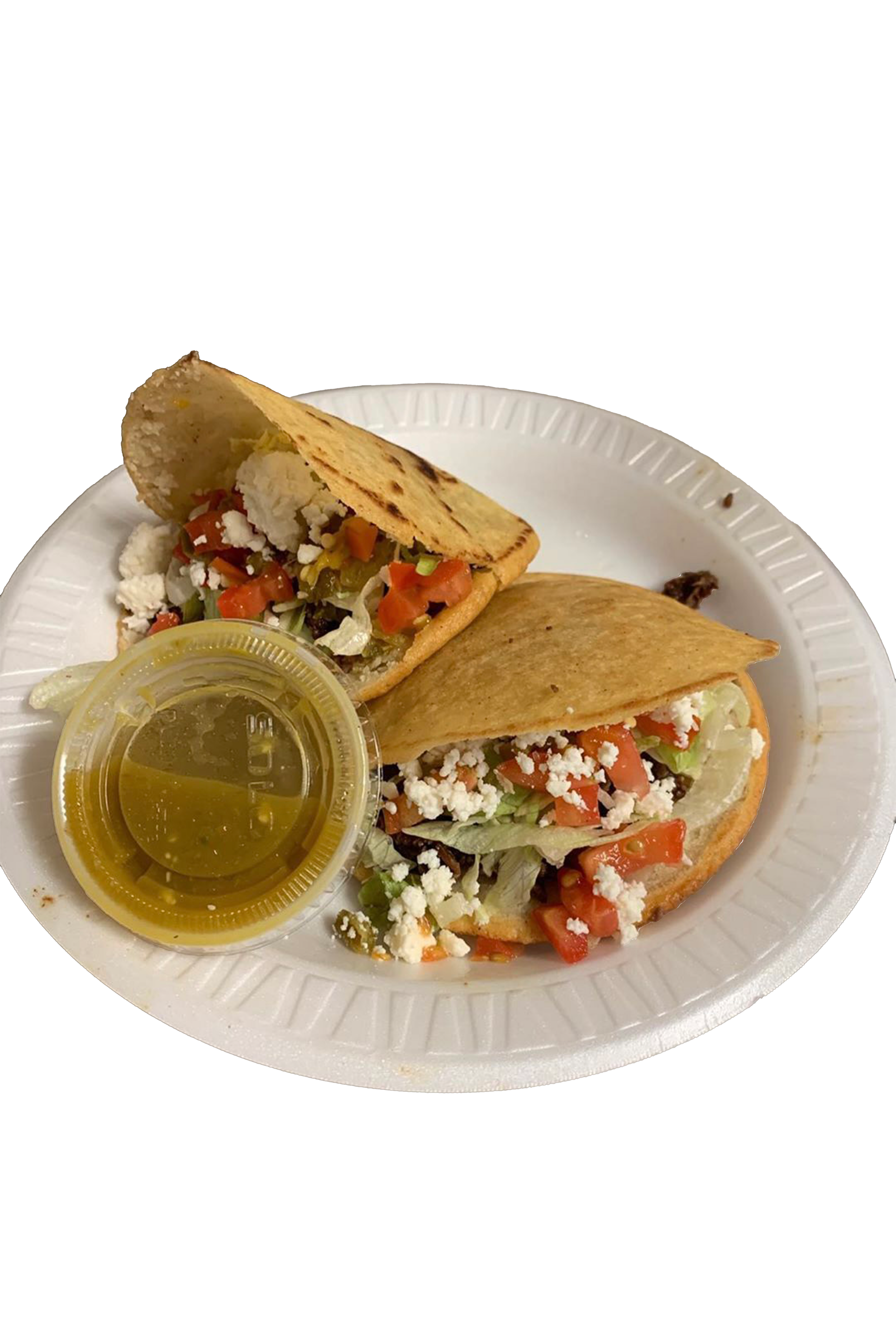 Two Tex-Mex style tacos filled with lettuce, chopped tomatoes, shredded cheese, and crumbled queso fresco, served on a white disposable plate with a small container of green salsa and a cup of yellow-green beverage.