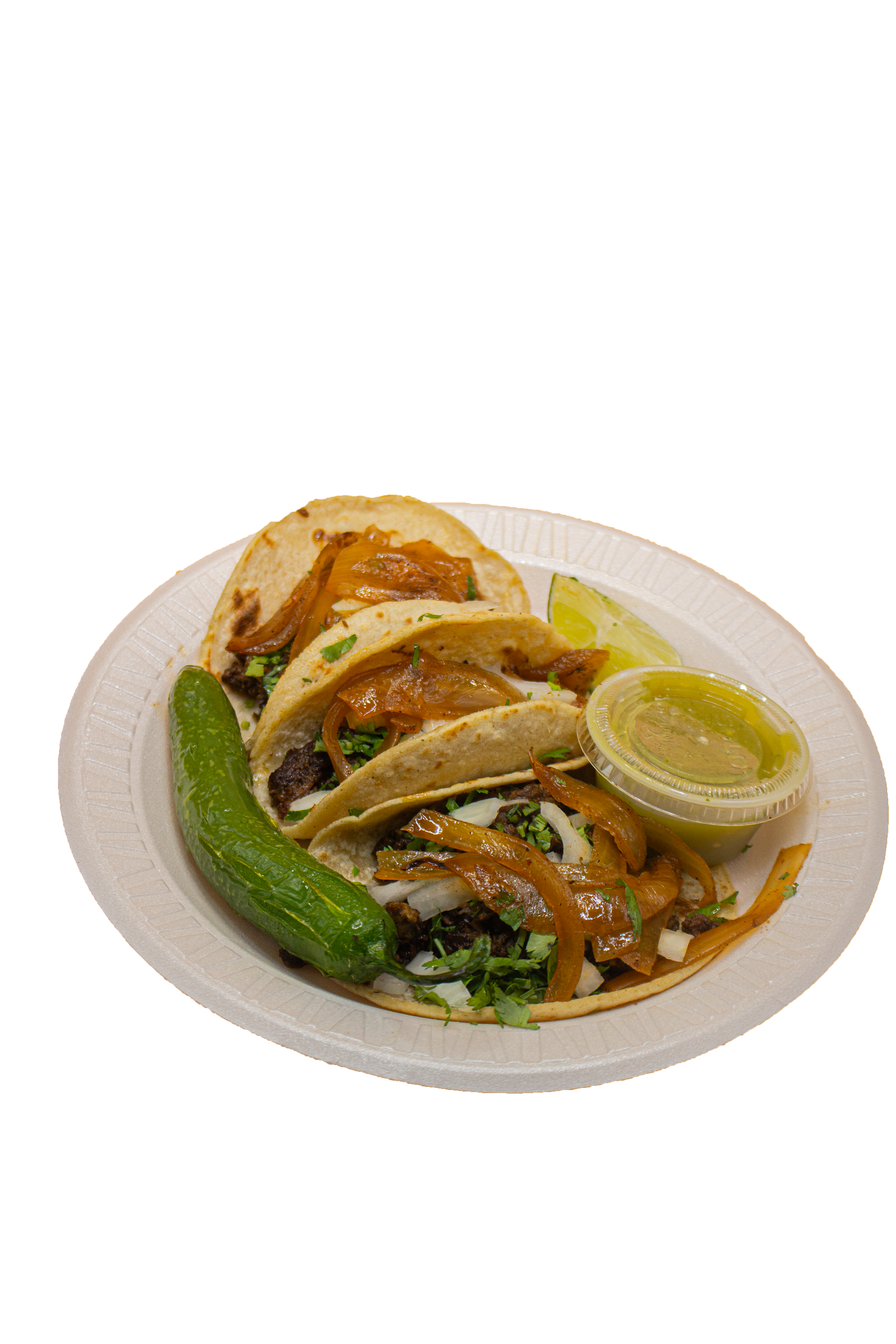 A white disposable plate with three tacos filled with meat, grilled onions, salsa, and chopped herbs, served with a lime wedge and a small cup of green sauce, on a wooden surface.