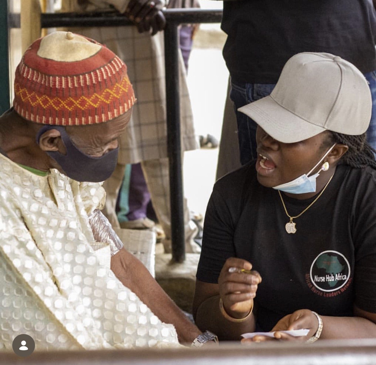 A woman from Nurse Hub Africa talking to an elderly man wearing a red patterned hat and face mask at a community health setting.