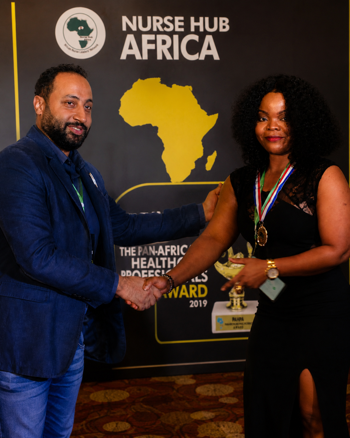 A nurse being recognised at the Nurse Hub Africa award ceremony, with a large banner in the background featuring a map of Africa and the event details "Pan-African Healthcare Professional Award" 2019