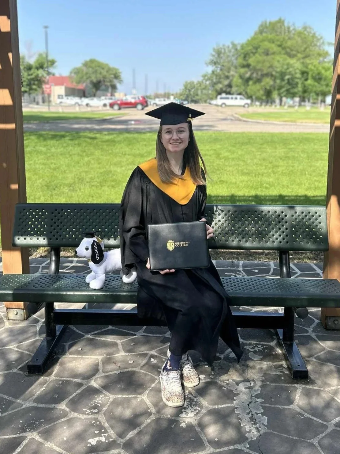 A young woman in a black graduation gown and cap sitting on a park bench outdoors with trees and a parking lot in the background, holding a diploma. There is a white plush dog with a neckband marked '2025'.