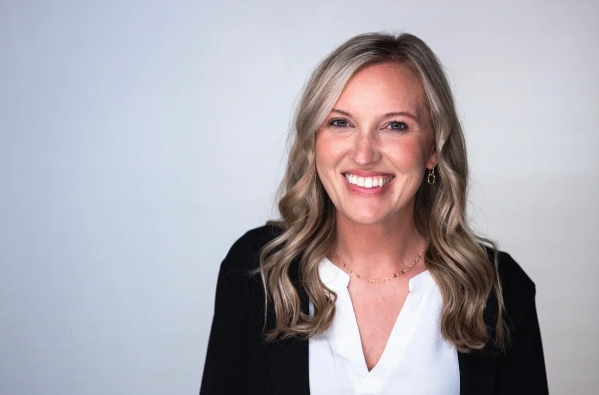A woman with long wavy blonde hair smiling at the camera, wearing a black blazer over a white top, gold earrings, and a delicate gold necklace. Katelin DeVore