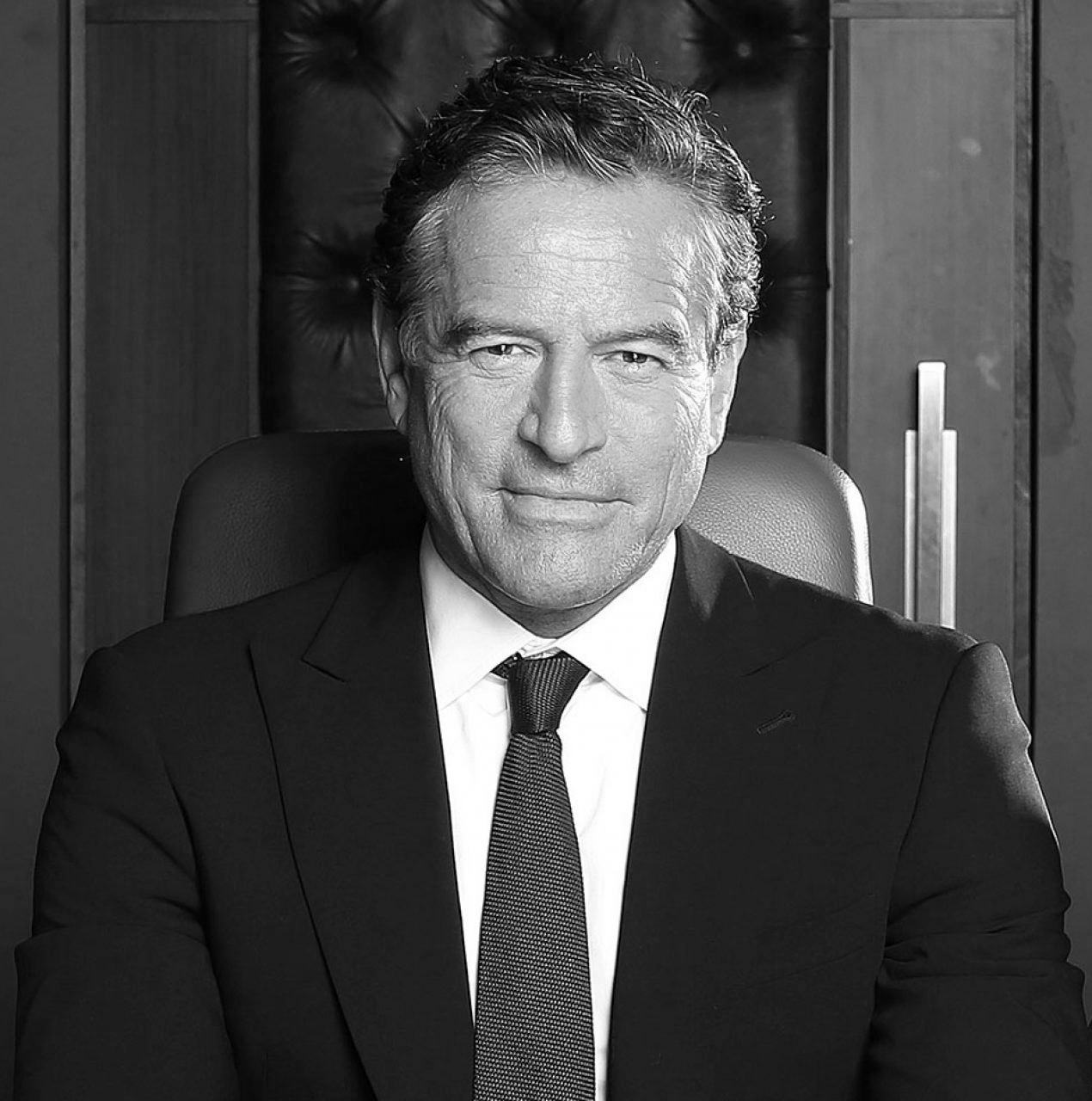 A black and white photo of a middle-aged man with dark hair, sitting in a leather chair in an office setting. He is wearing a dark suit, white shirt, and a tie, and has a confident expression.