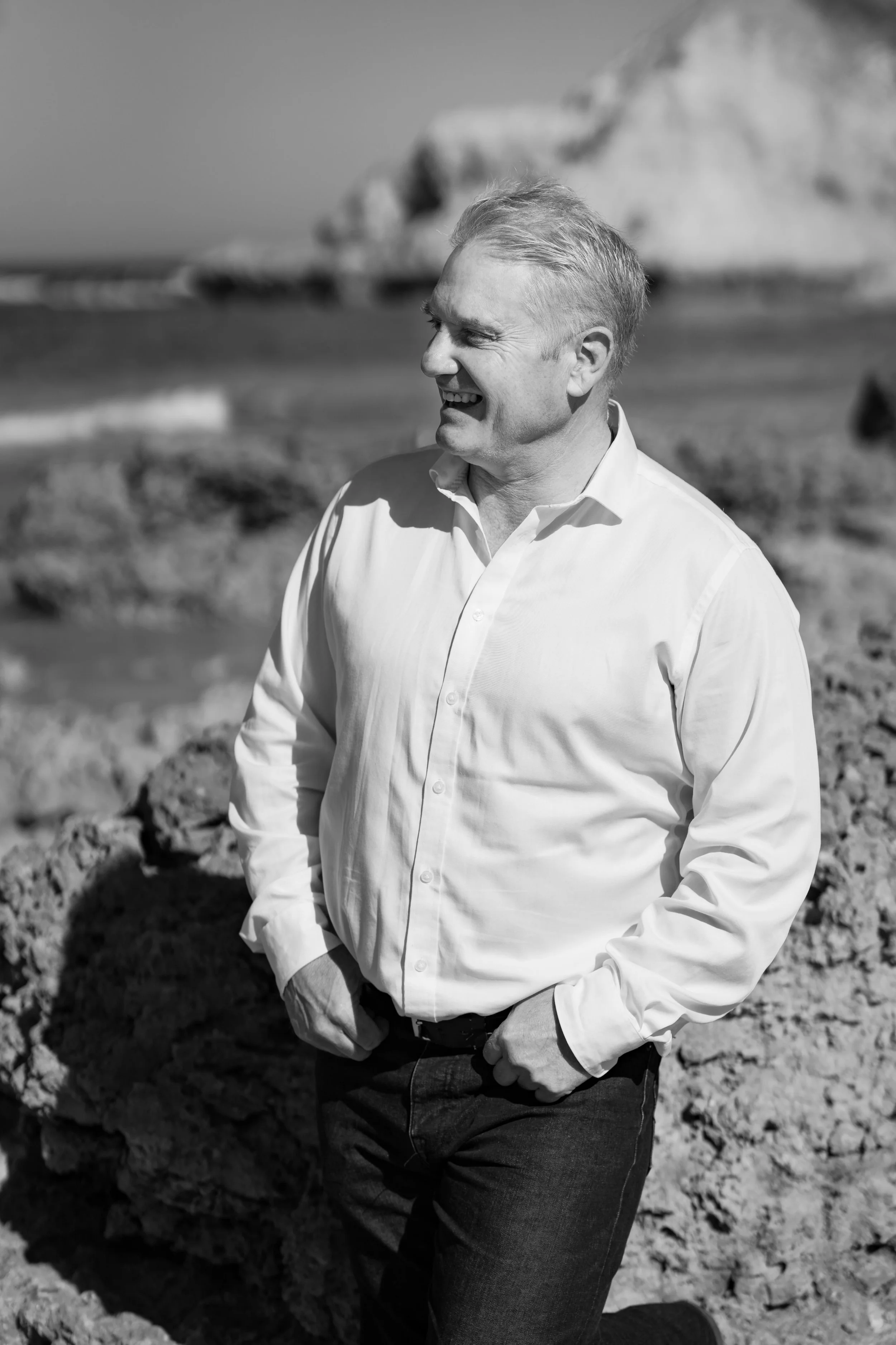A man in a white shirt and jeans smiling while looking to the side at the rocky beach with cliffs in the background.