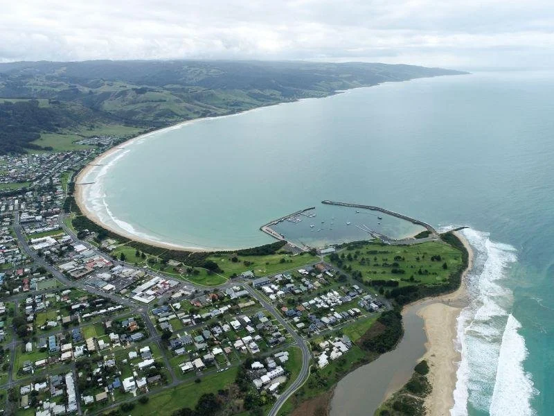 Aerial view of a coastal town with a crescent-shaped beach, marina, and green park near the shore, with ocean waves crashing on the beach.