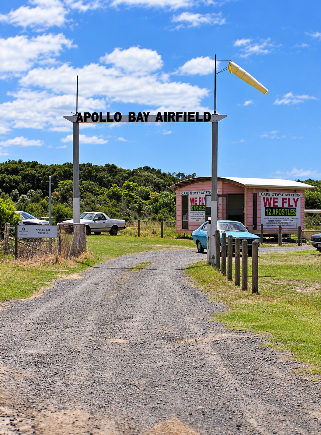 Entrance sign for Apollo Bay Airfield with a gravel road leading to a pink building, parked cars, and a windsock against a partly cloudy sky and green foliage background.