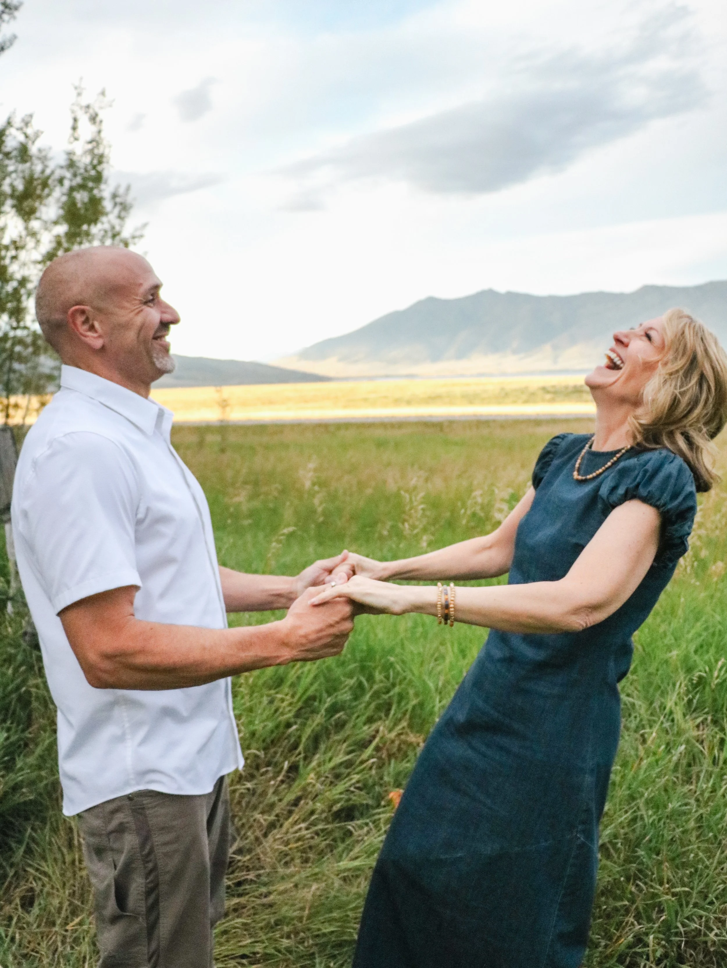 A man and woman holding hands and smiling in a grassy field with mountains and cloudy sky in the background.