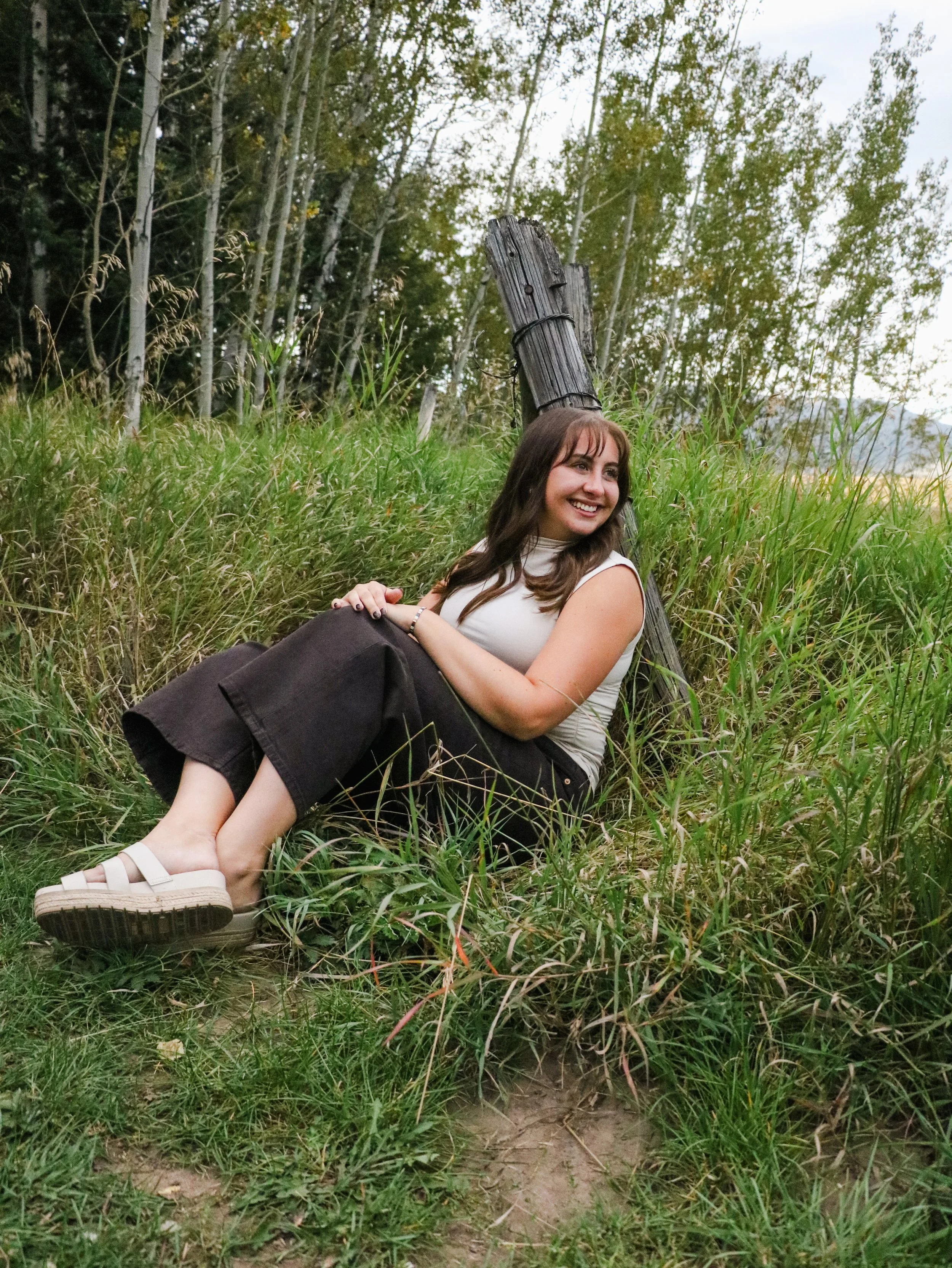 A woman with brown hair and a white sleeveless top sitting on grass and leaning against a wooden post in a natural outdoor setting with trees in the background, smiling and looking to her right.