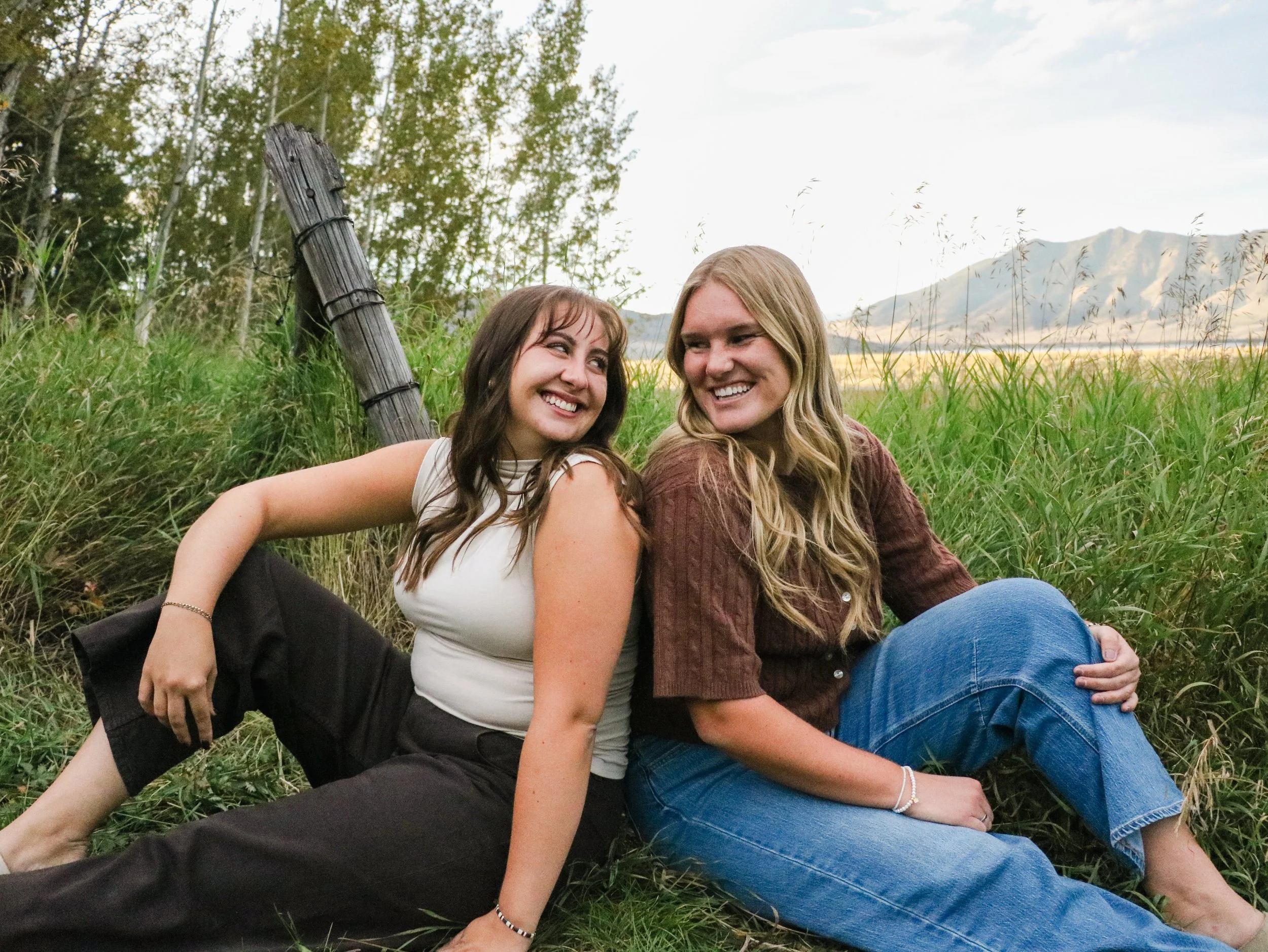 Two young women smiling and sitting close together in a grassy outdoor setting with trees and mountains in the background.