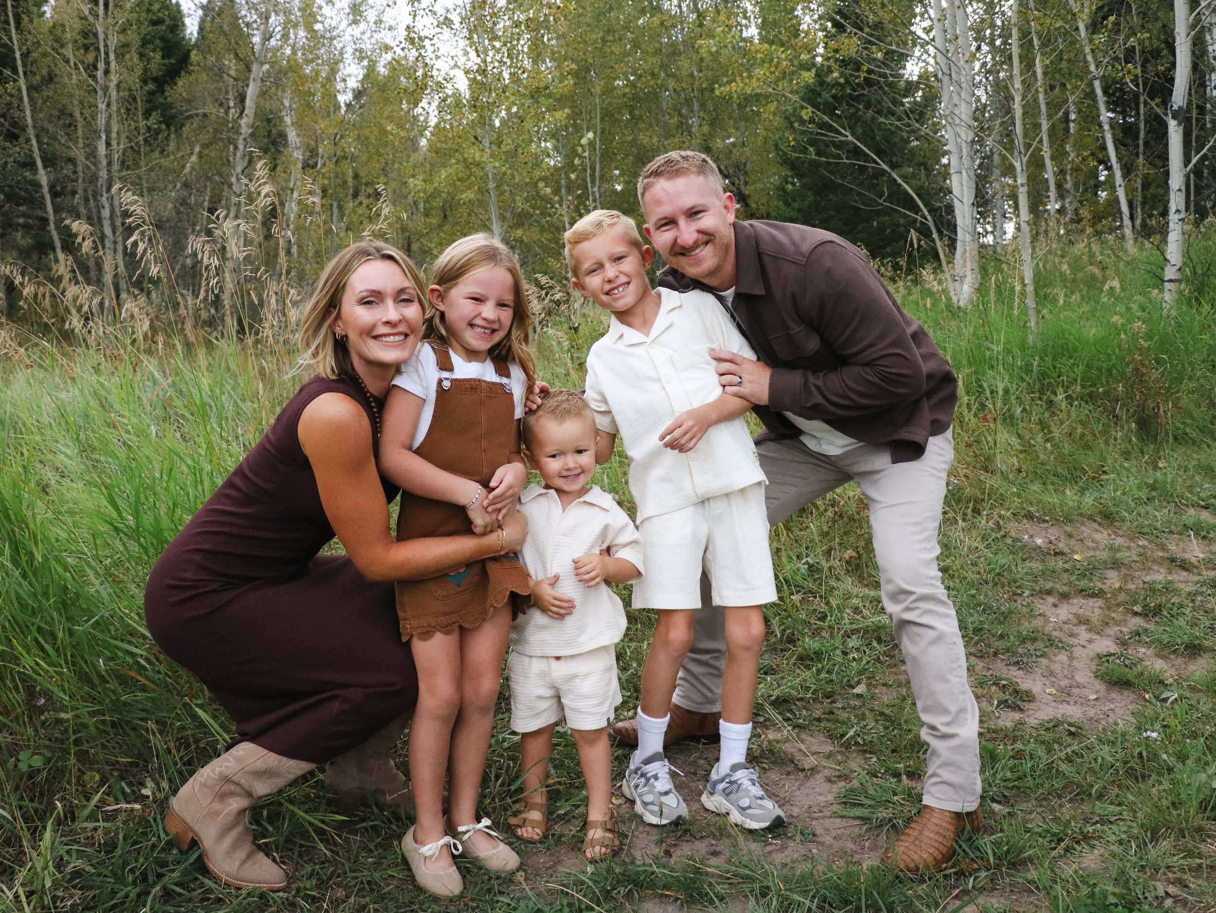 A family of six outdoors in a grassy area with trees in the background, smiling and embracing each other.