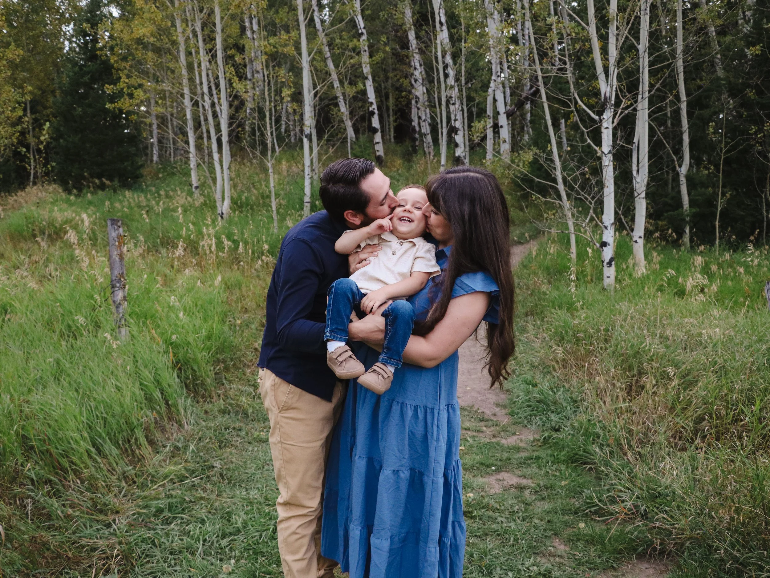 A family of three, a man, a woman, and a young child, standing outdoors in a forest clearing, smiling and kissing each other, with trees in the background.