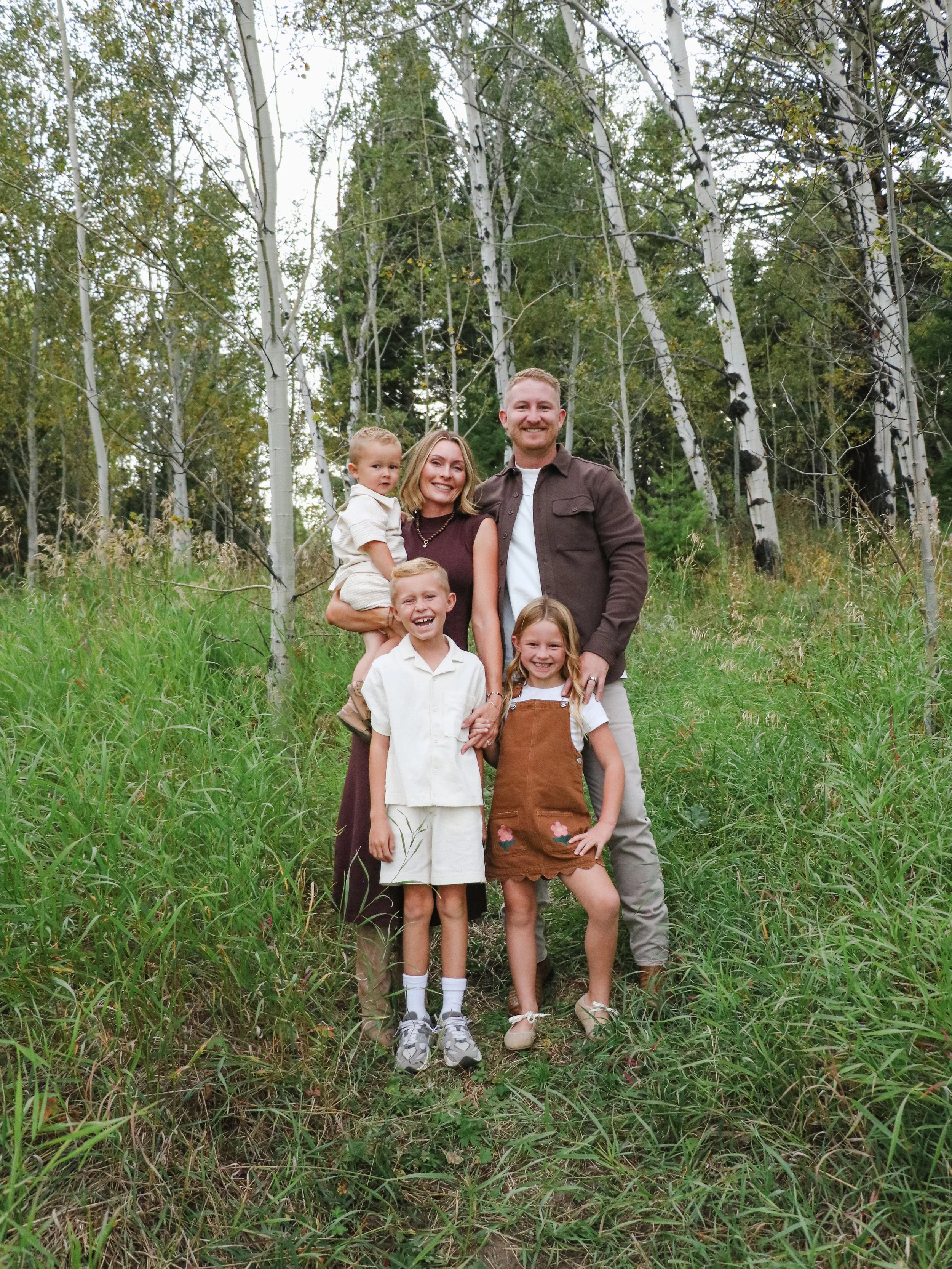 A family of six, including two adults and four children, posing outdoors in a forest of tall, white-barked trees with green foliage, on a grassy area.