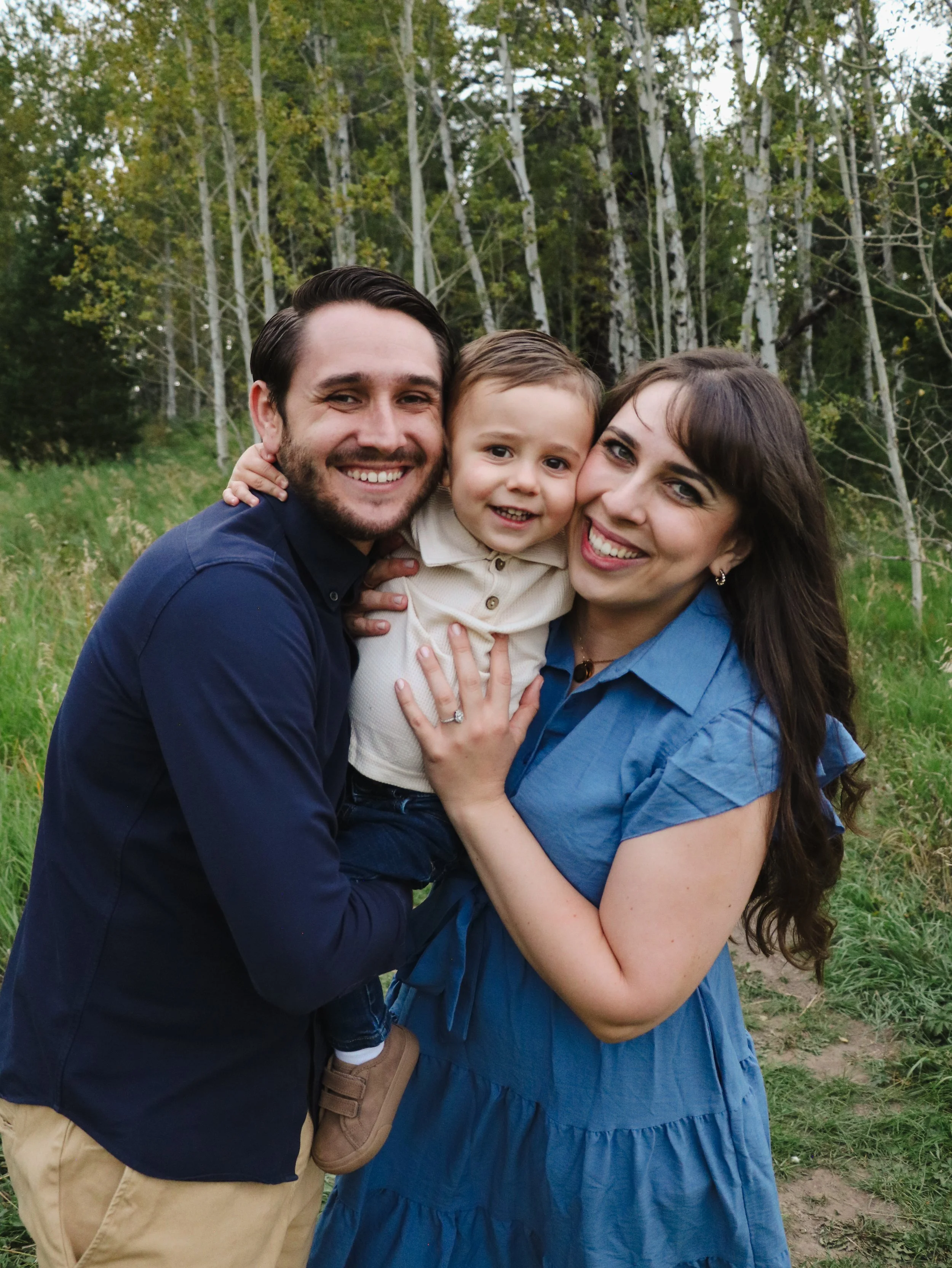 A smiling family of three having a hug outdoors, standing in front of a forested area with trees and green grass.