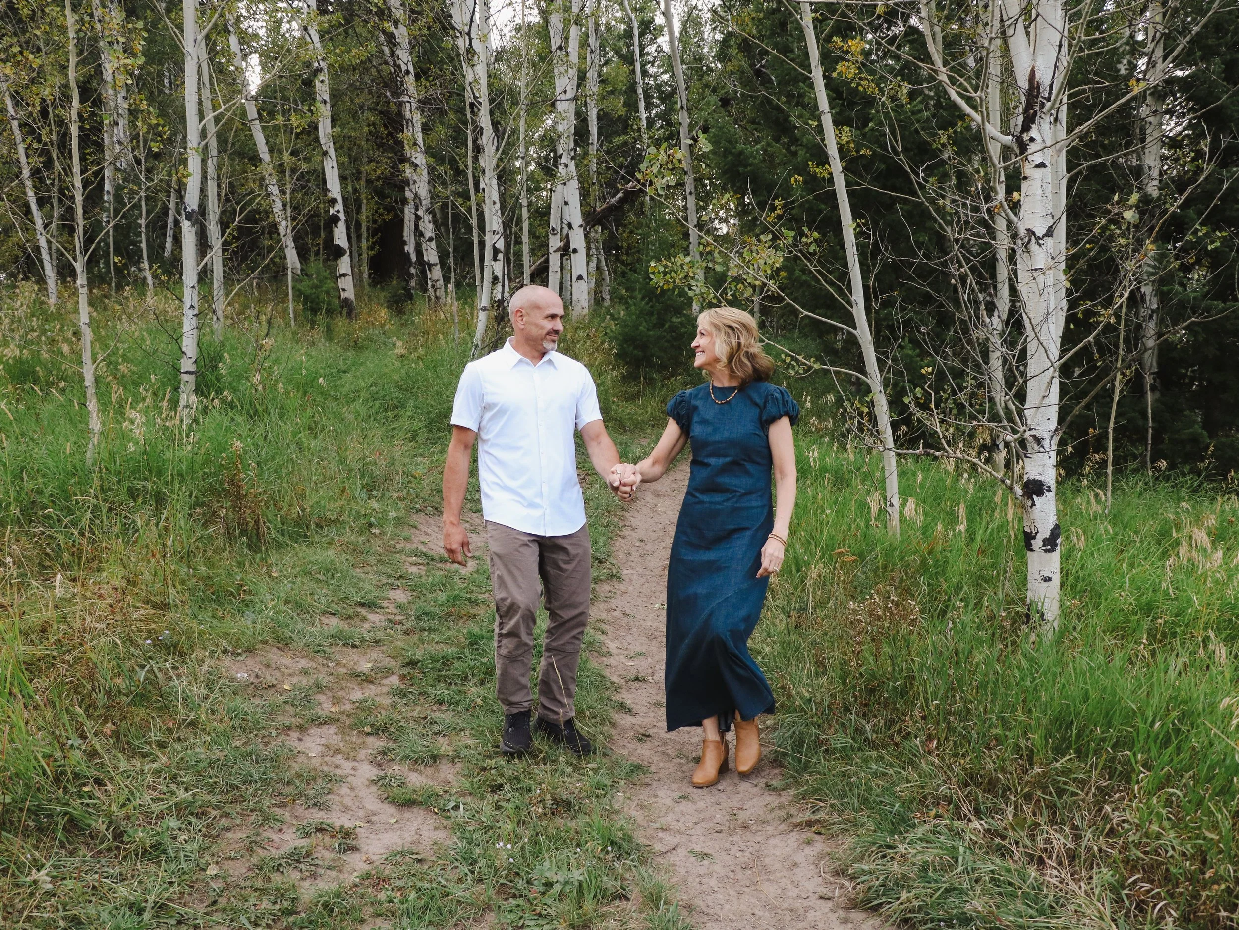 A couple walking hand-in-hand on a dirt trail through a forest of tall white-barked trees, with green grass and foliage around them. The man has a bald head and is wearing a white shirt and light-colored pants. The woman has shoulder-length blonde ha