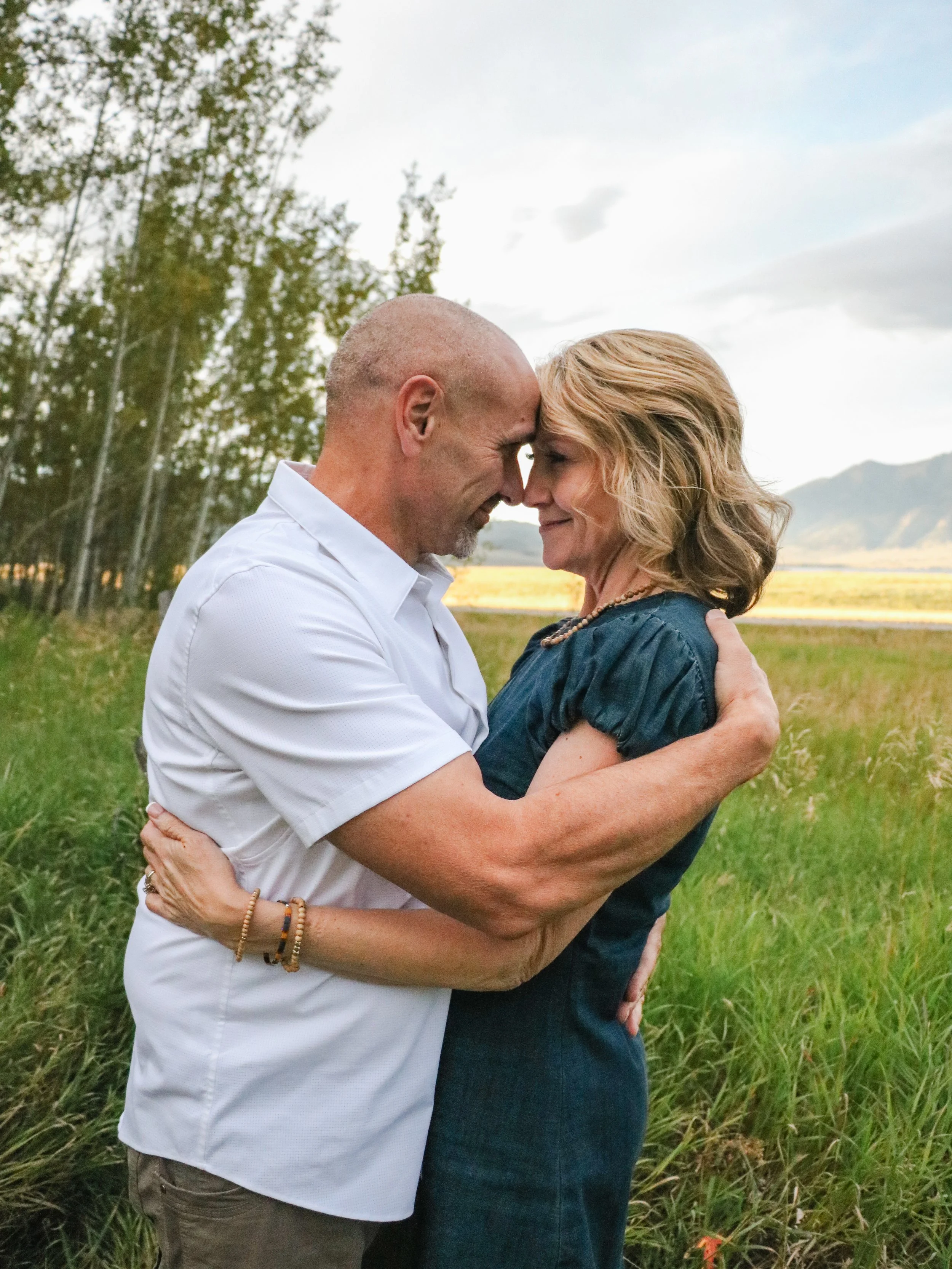 A middle-aged man and woman with light skin embrace outdoors in a grassy field, touching foreheads, with trees, mountains, and a cloudy sky in the background.