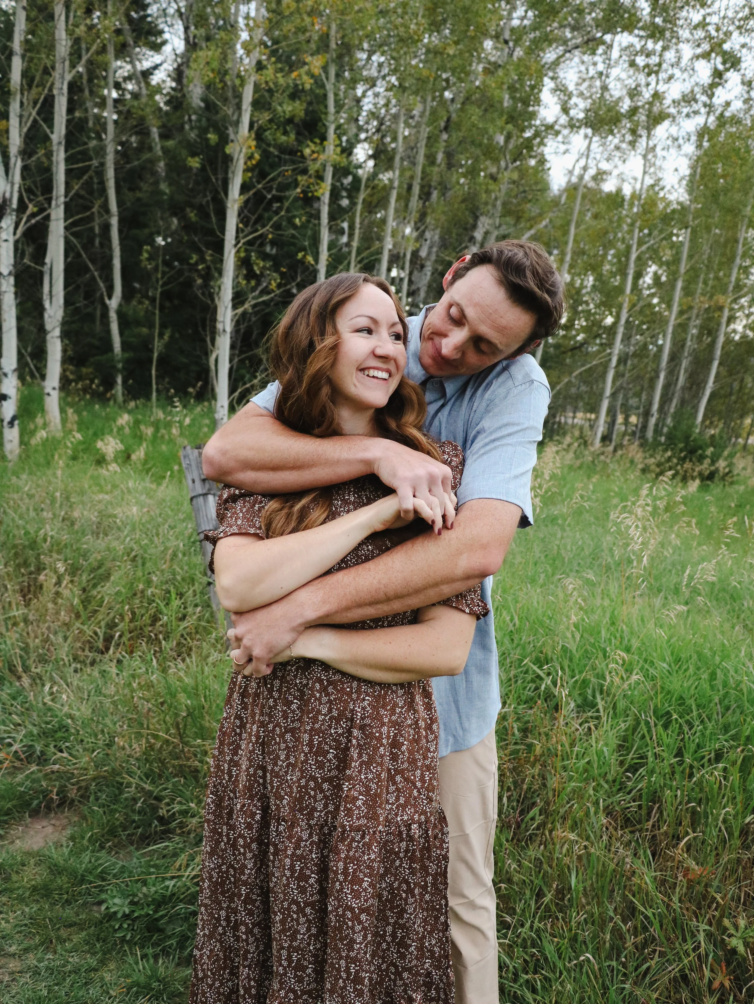 A young woman and a man are outdoors in a grassy area with trees in the background. The woman, with long hair and wearing a brown floral dress, is smiling and embracing the man. The man, with short hair and wearing a light blue shirt, is hugging the 