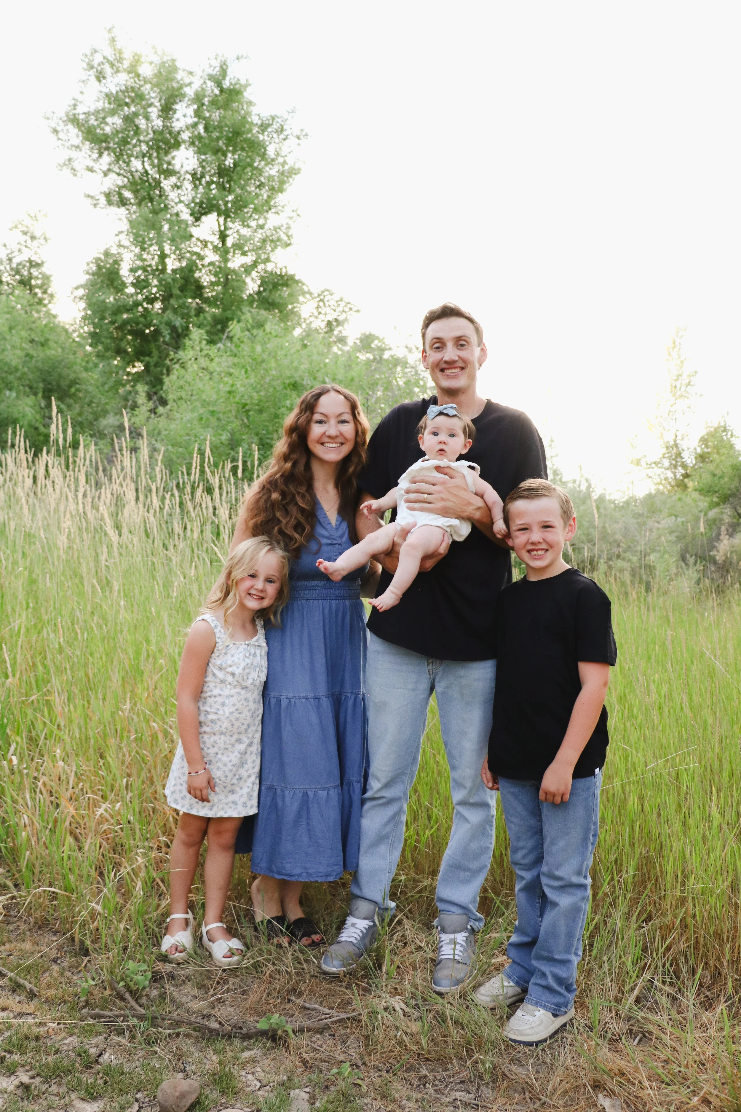 Family of five standing outdoors on a grassy field with trees in the background, four children and one adult woman, all smiling.