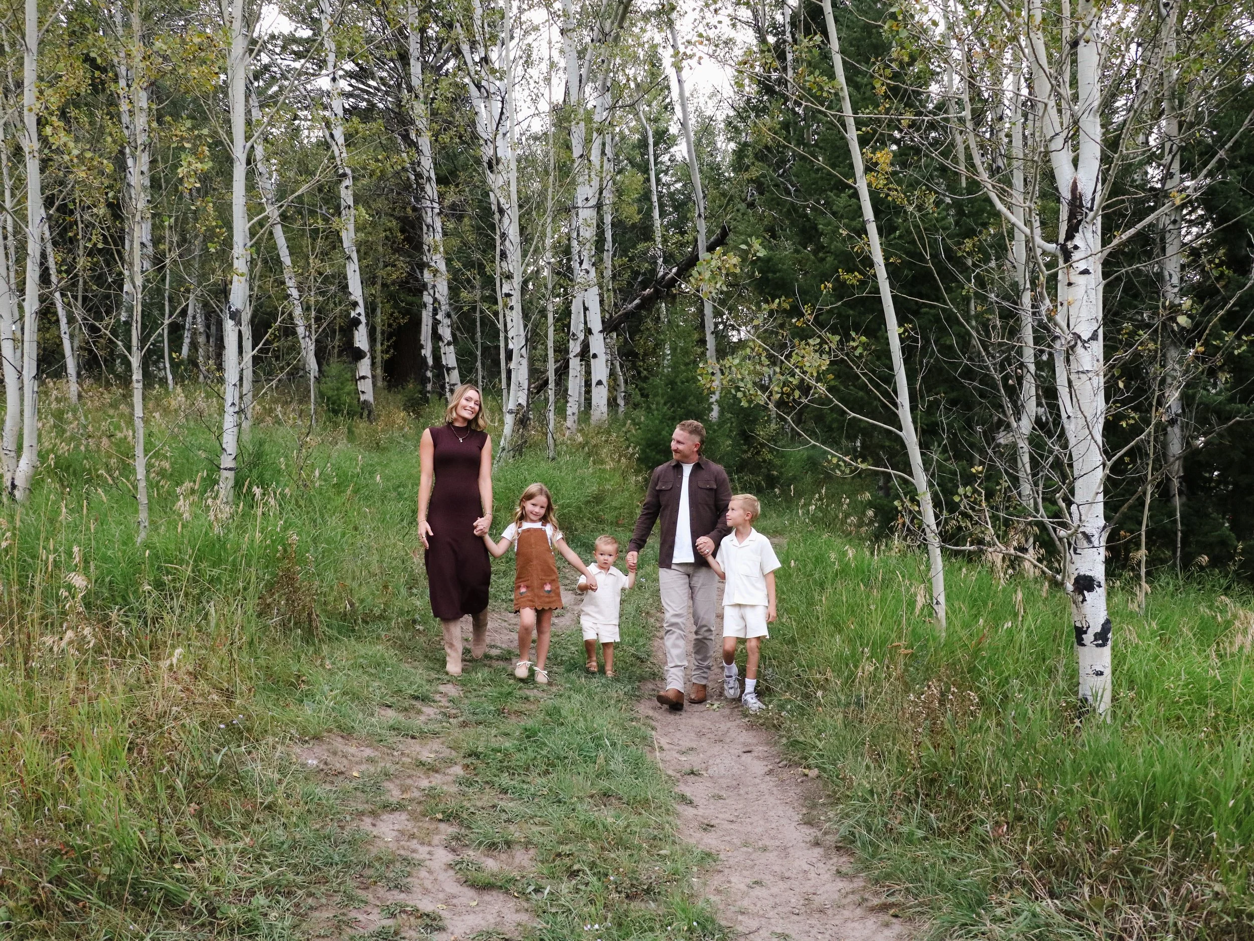 A family of six walking along a dirt trail through a forest of white-barked aspen trees with green foliage, some turning yellow. The family includes two women, three children, and a man, all smiling and holding hands.