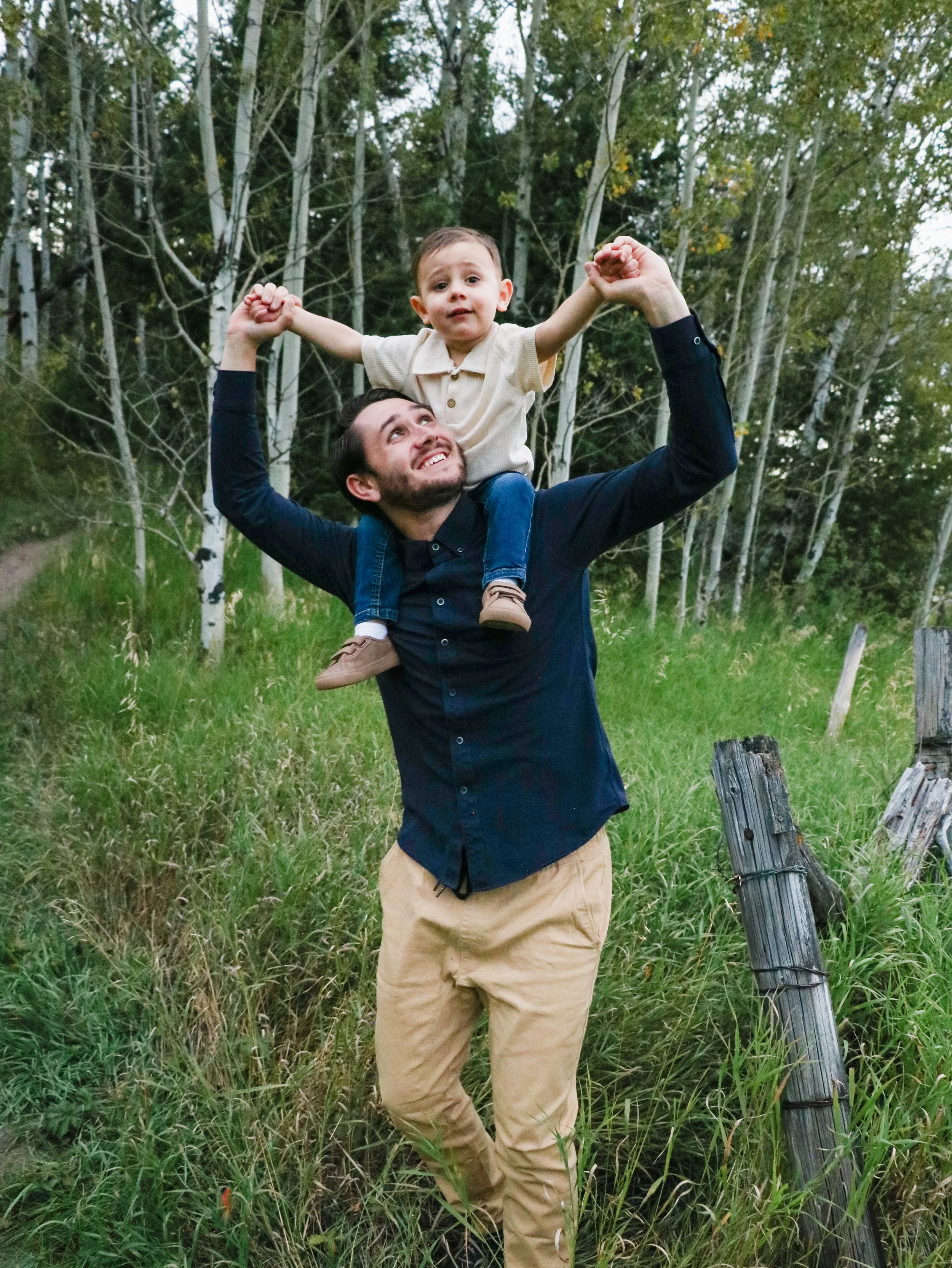 A man is holding a young boy on his shoulders with a forest of green trees in the background.