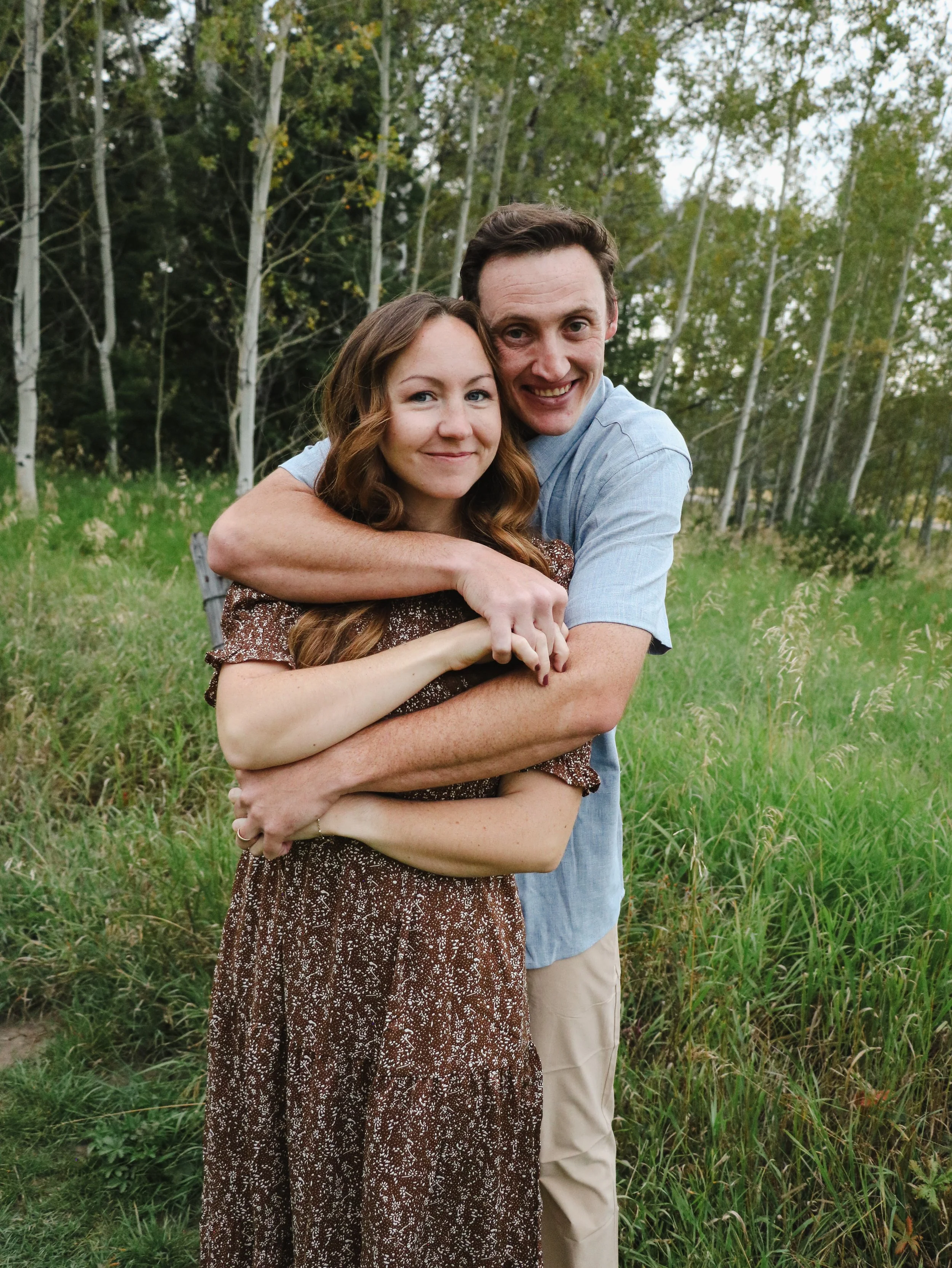 A couple hugging outdoors in a grassy field with trees in the background.
