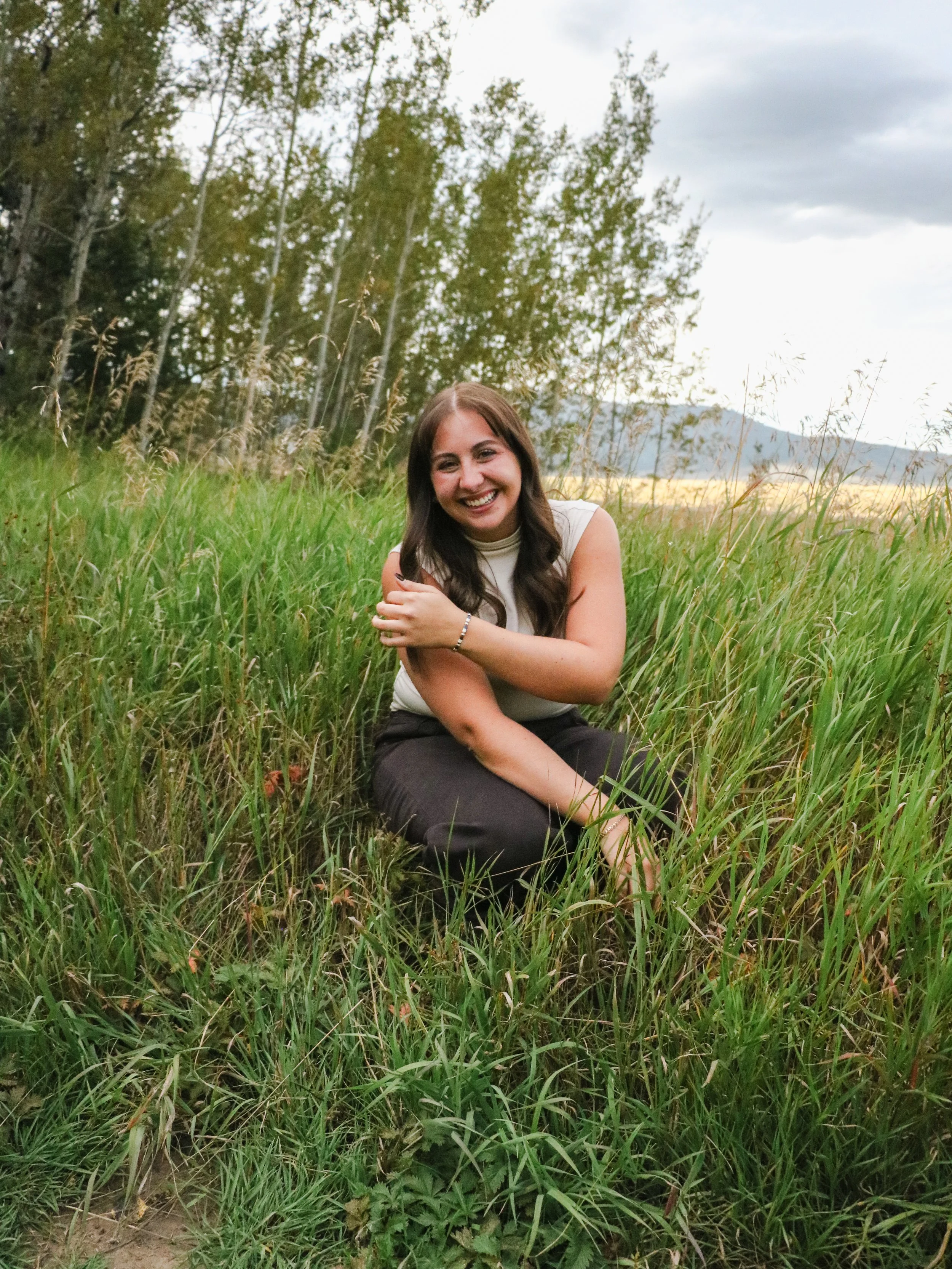 Smiling woman with long brown hair sitting in green grass during daytime, surrounded by trees with cloudy sky in the background.